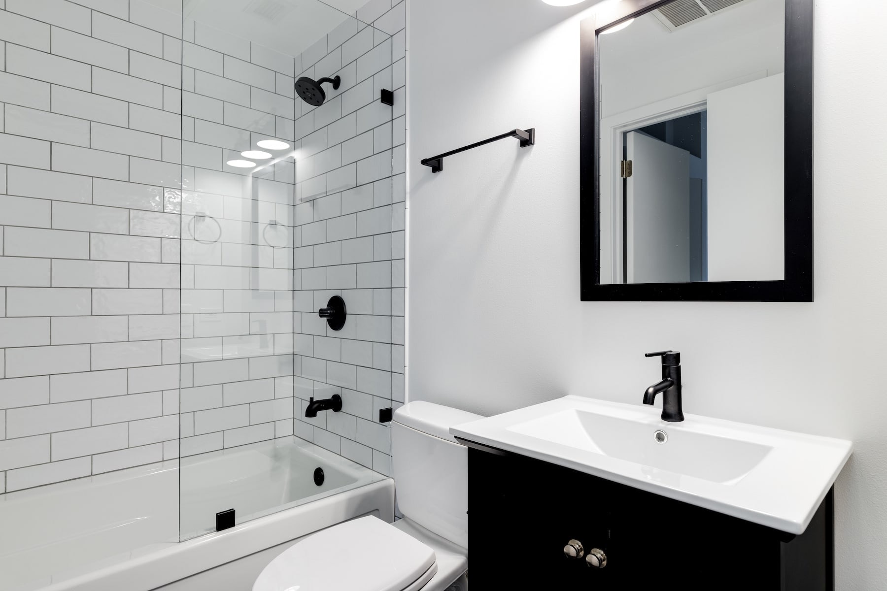 White metro tiles on the wall behind a bath with a shower above it, beside a vanity unit and a black-framed mirror.