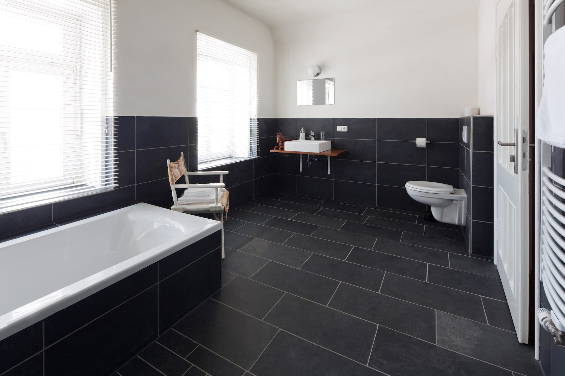 A bathroom floor laid with large black tiles, with a built-in bath and a wall-hung toilet and basin.