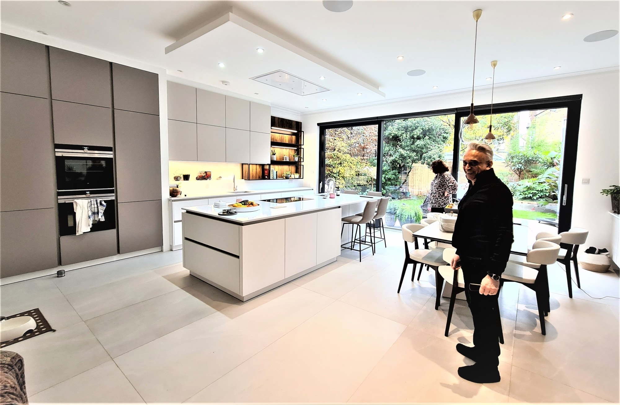 Open-plan contemporary kitchen with white handleless island and matching base units, tall matt grey cabinetry with built-in ovens, pale worktop, and light grey tiled floor. Pendant lights overhead and two people in the room.