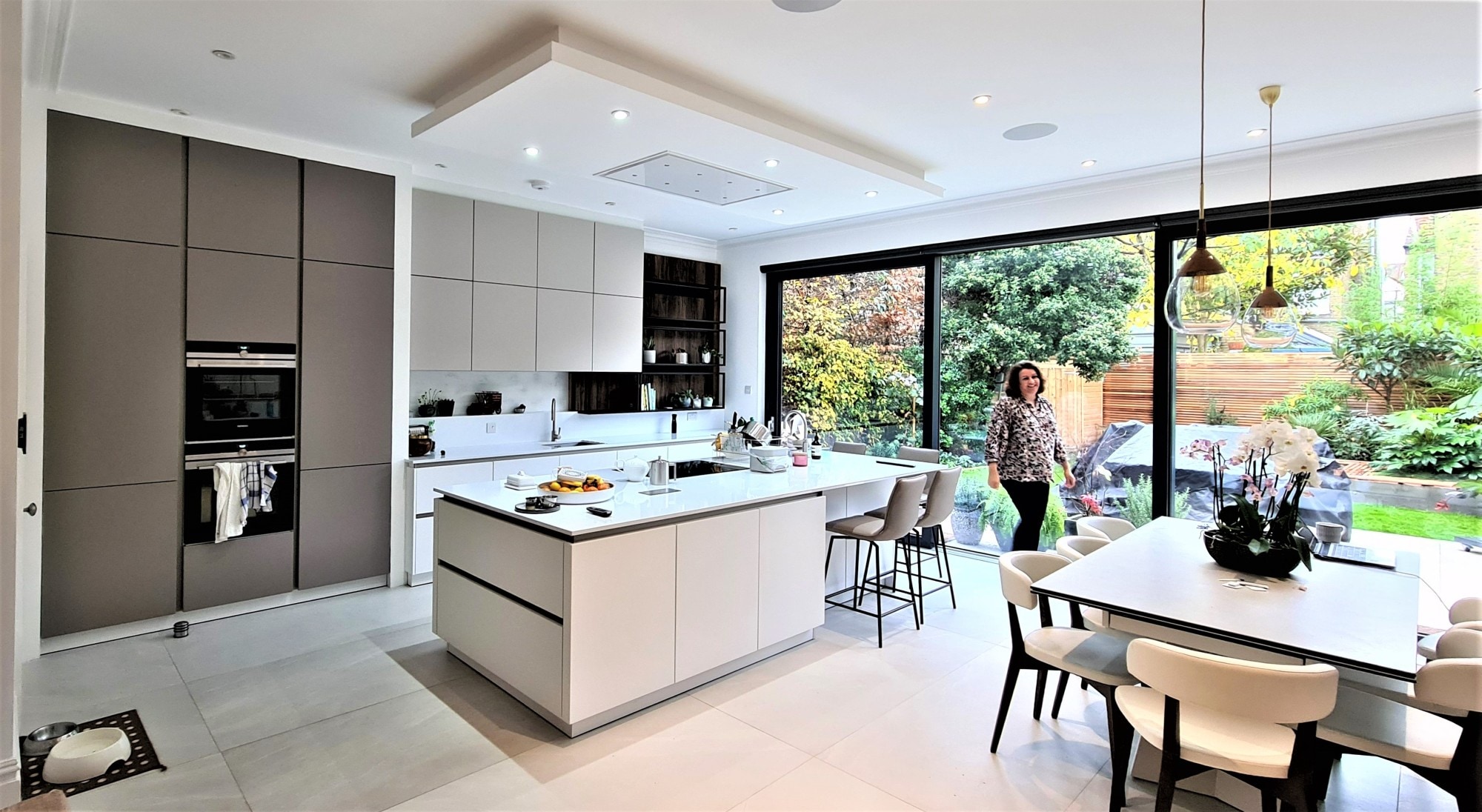 Open-plan contemporary handleless kitchen with a large white island, white and warm grey cabinetry, integrated double ovens, and white worktops.