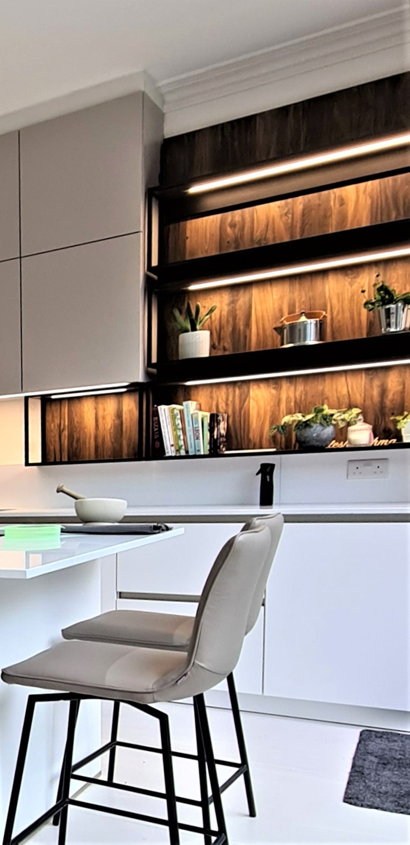 Contemporary kitchen with matt light-grey handleless cabinetry, a pale worktop and an overhanging breakfast bar with a taupe bar stool. Dark metal-framed open shelves with warm wood-effect back panels and integrated LED strip lighting display books, plants and cookware.