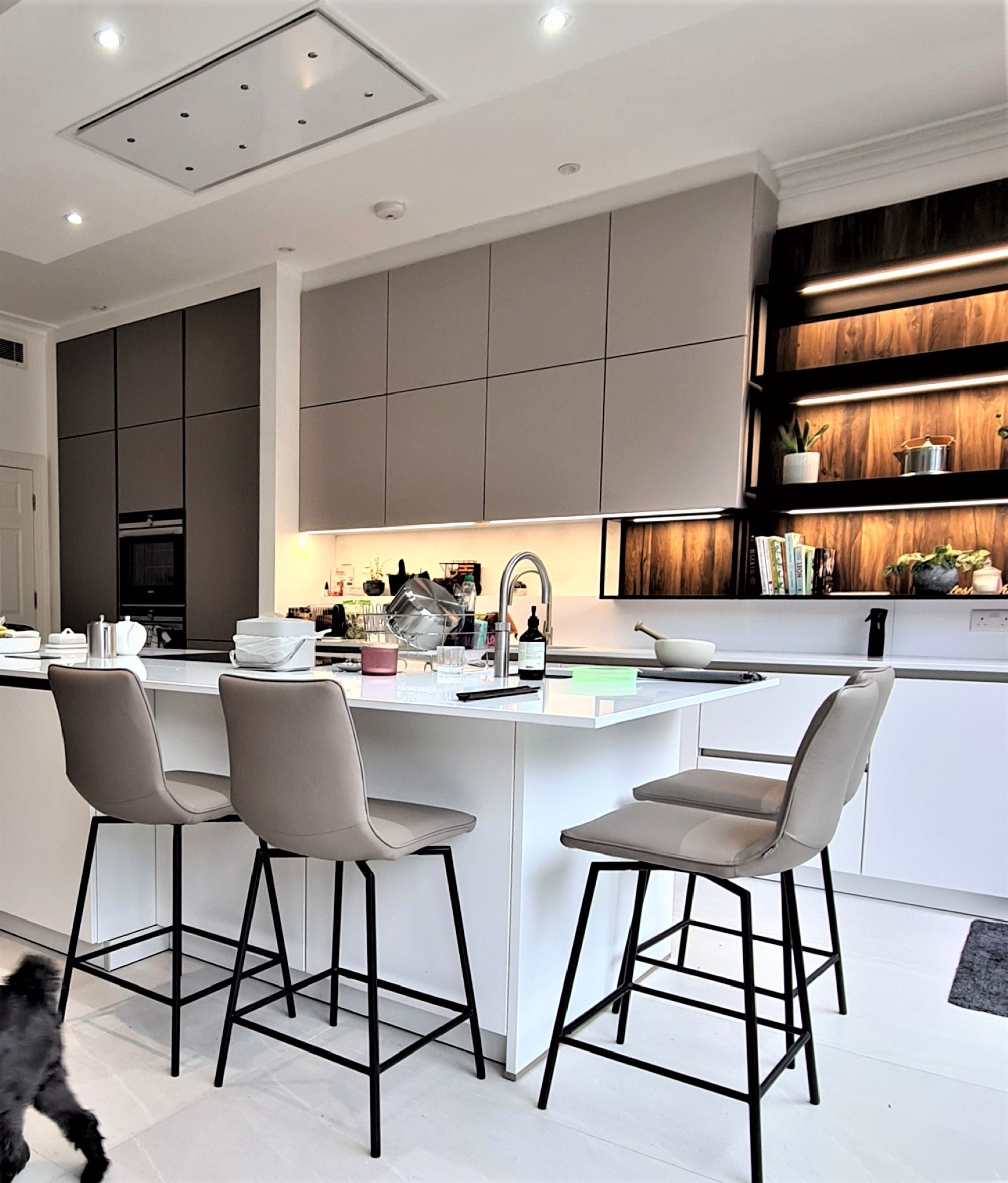 Contemporary handleless kitchen with a white island and white worktop, grey wall cabinetry, built-in oven housing, and open wood-effect shelving with warm LED lighting. Three upholstered bar stools sit at the island, with an inset sink and chrome tap on the worktop.