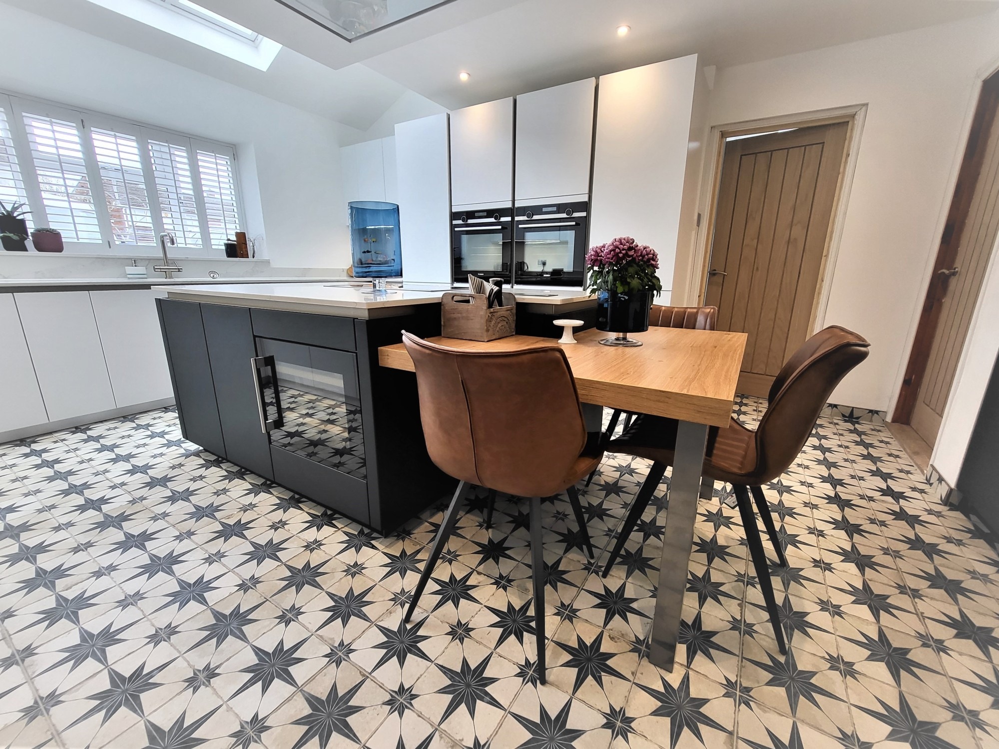 Contemporary german kitchen with handleless white and dark grey cabinetry, a large island with a pale stone-effect worktop, integrated double ovens, and a built-in dining table with brown upholstered chairs on patterned tile flooring.