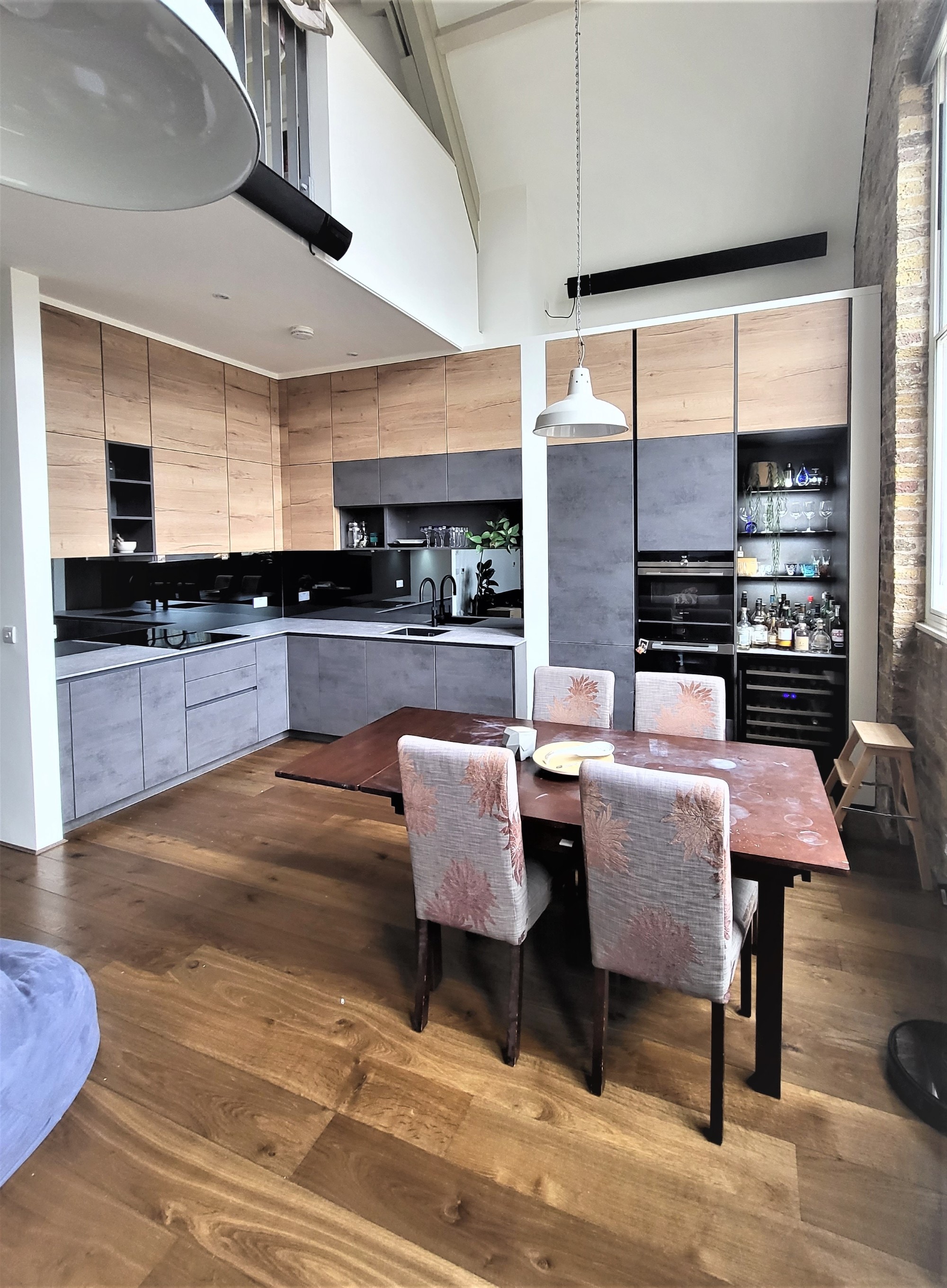 Modern German kitchen with handleless wood-effect and grey concrete-effect cabinetry, black worktops and splashback, built-in ovens and wine fridge, and a dining table beside tall industrial-style windows and wood flooring.