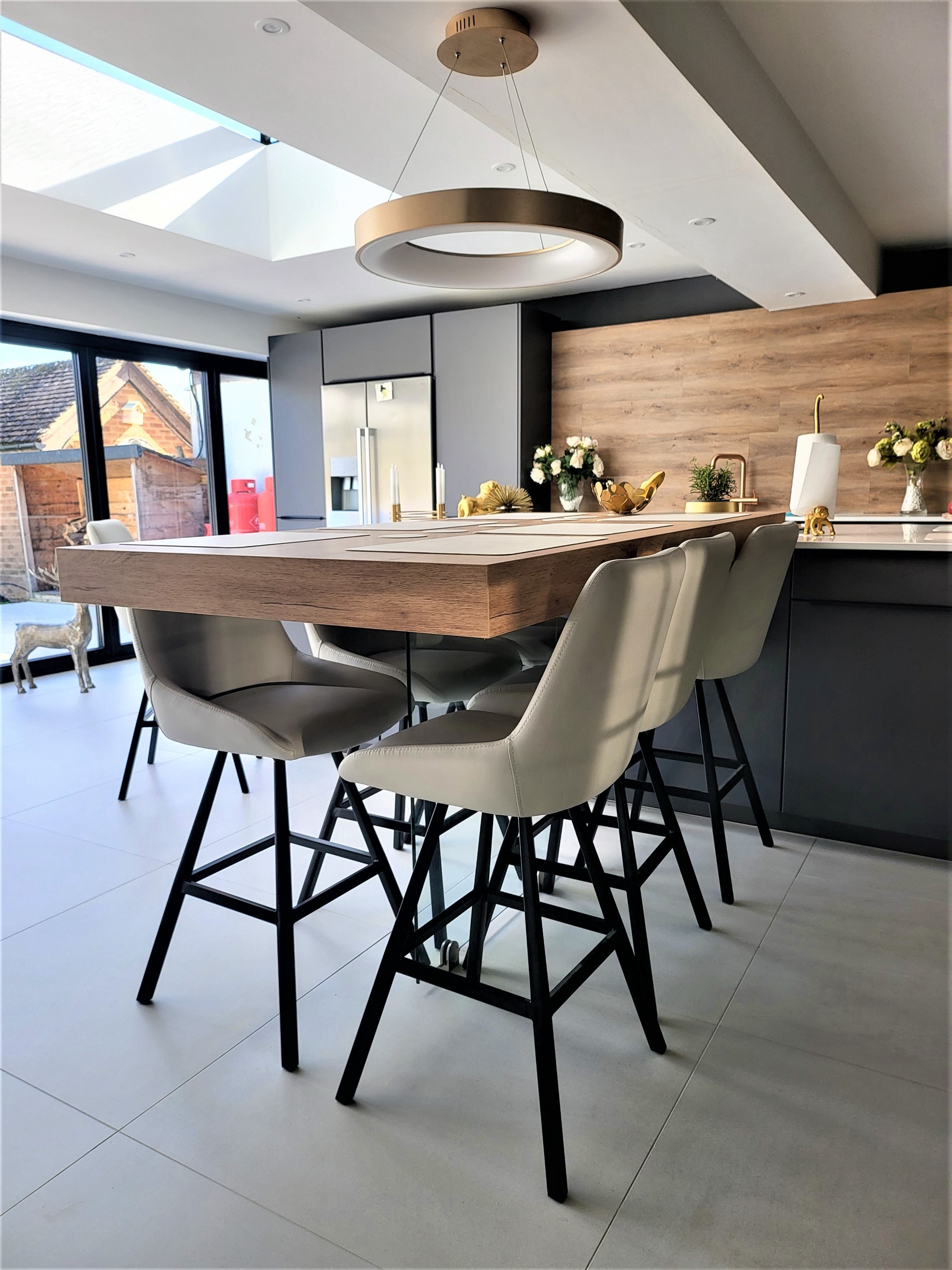 Contemporary open-plan kitchen with matt dark grey handleless cabinetry, a large island with wood-effect breakfast bar overhang, pale stone-effect worktops, bar stools, brass-toned tap, wood-effect splashback, and a circular pendant light under a roof skylight.