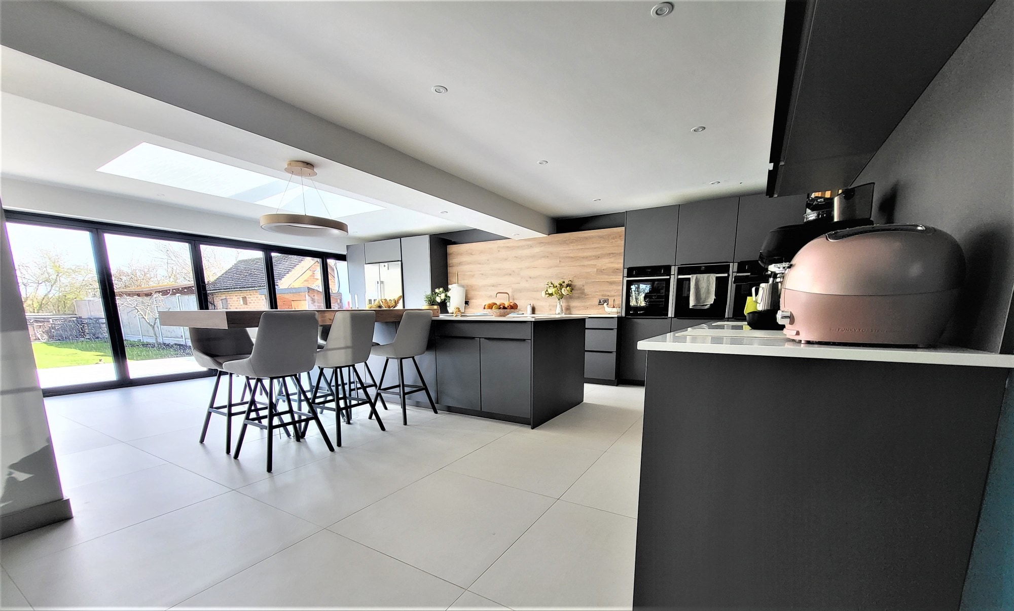 Open-plan contemporary kitchen with matt dark grey handleless cabinetry, a large island with a white worktop and breakfast bar seating, integrated double ovens, wood-effect splashback, and wide sliding glass doors to the garden.