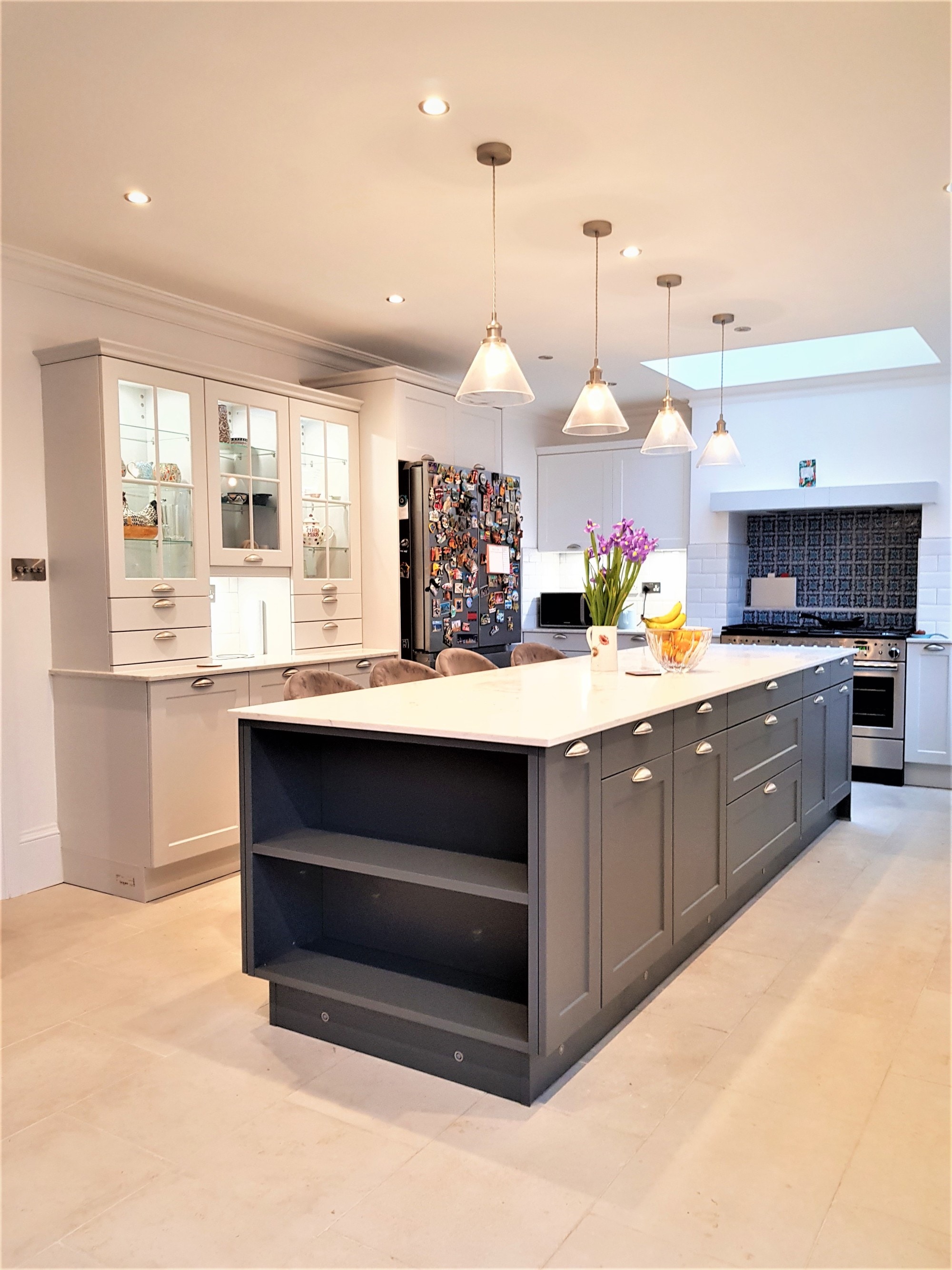 Bespoke shaker kitchen with a large dark grey island and open end shelving, a white stone-effect worktop, cream cabinetry with glazed display cupboards, white tiled splashback, and pendant lighting above the island.