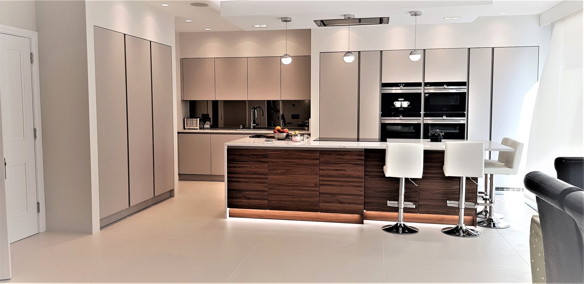 Bright contemporary kitchen with handleless light taupe cabinetry, a dark wood-effect island with white waterfall-style worktop, integrated oven stack, black tap at the sink, pendant lighting and a breakfast bar with white stools on large light floor tiles.