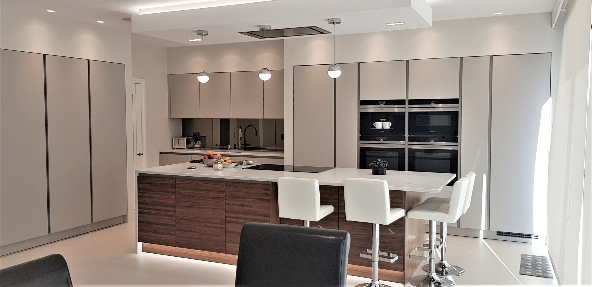 Large contemporary German kitchen with handleless light grey tall cabinetry, a walnut-effect island, and a white stone-effect worktop with integrated hob and breakfast bar seating. Wall units sit above a sink run with a dark tap and reflective splashback, and a bank of built-in ovens is set into the tall units.