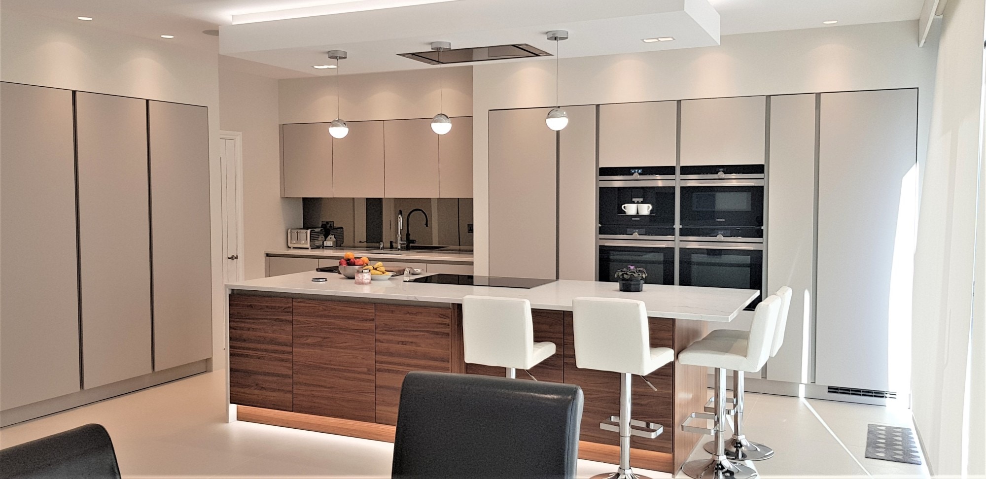 Bright contemporary German kitchen with handleless light-grey tall units, a large white island with wood-effect base, integrated hob and sink run, built-in ovens and three white bar stools under pendant lights.