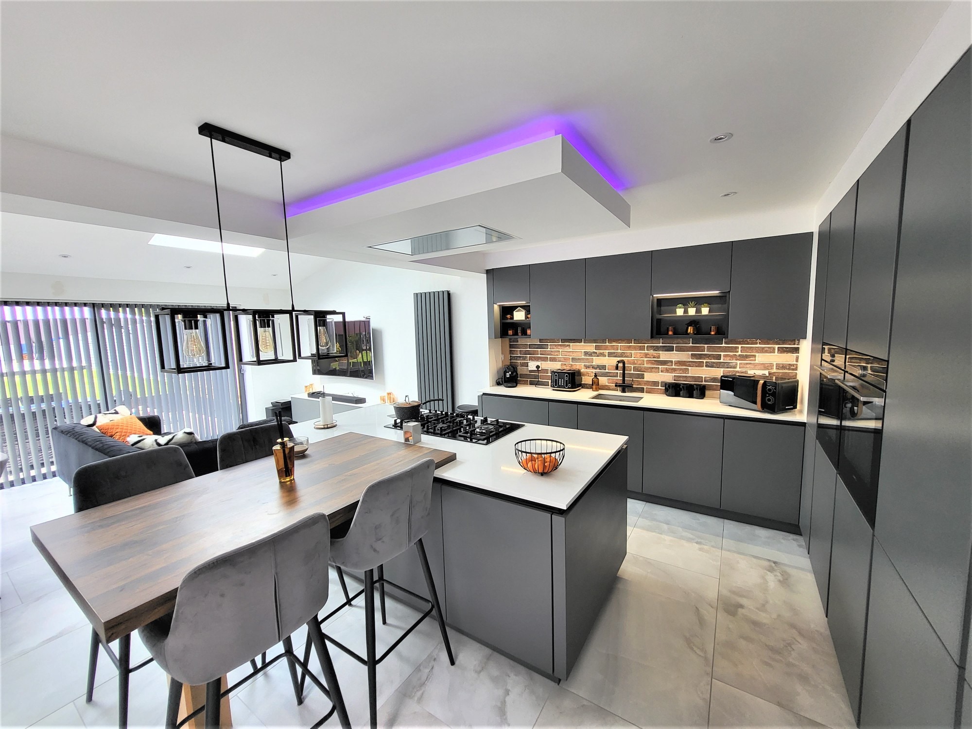 Open-plan German kitchen with matt dark grey handleless cabinetry, a white waterfall island with a black gas hob, brick-effect splashback, integrated ovens in a tall unit, and a wood-effect breakfast bar with upholstered stools. Large light grey floor tiles, black framed pendant lights, and a ceiling feature with coloured LED lighting are visible, with a lounge area beyond.