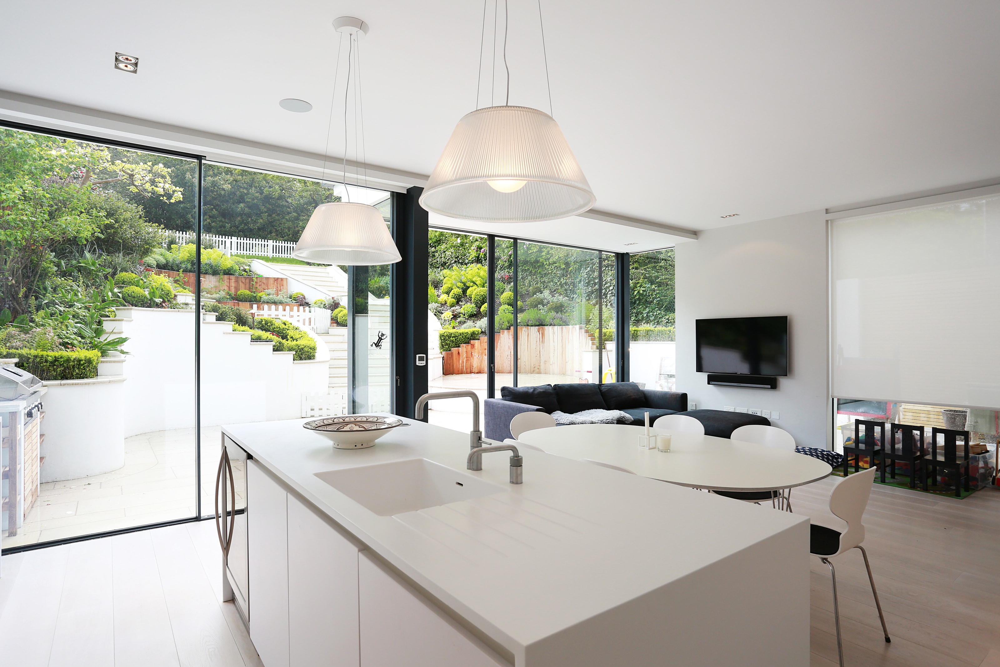 Open-plan Hacker kitchen in polar white with a clean, handleless island, integrated sink and chrome tap, opening to a dining area and lounge with full-height sliding glass doors to the garden.