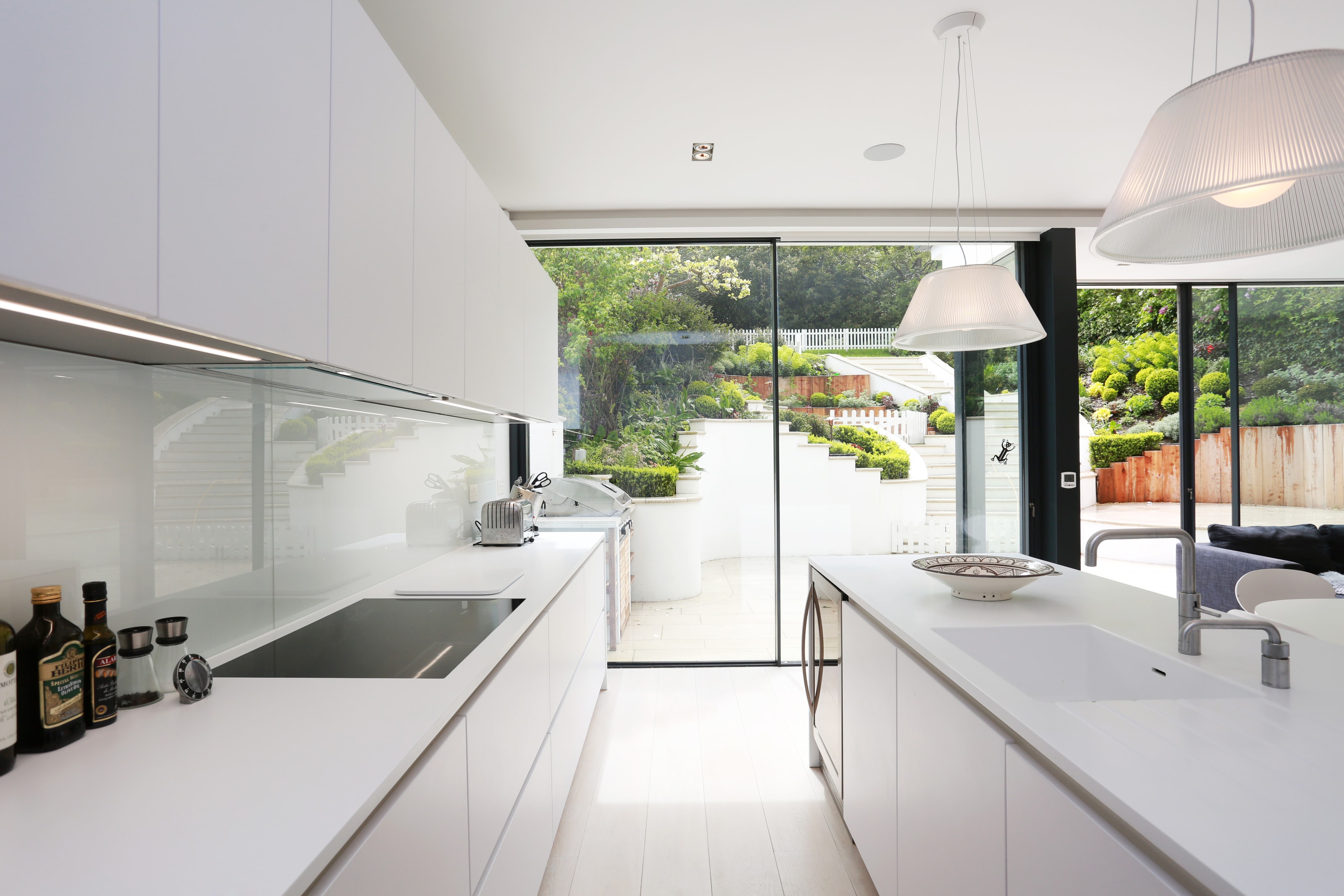 Clean white Hacker kitchen with polar white handleless cabinetry, a slim white worktop and glass splashback, plus an island with integrated sink facing large sliding garden doors.