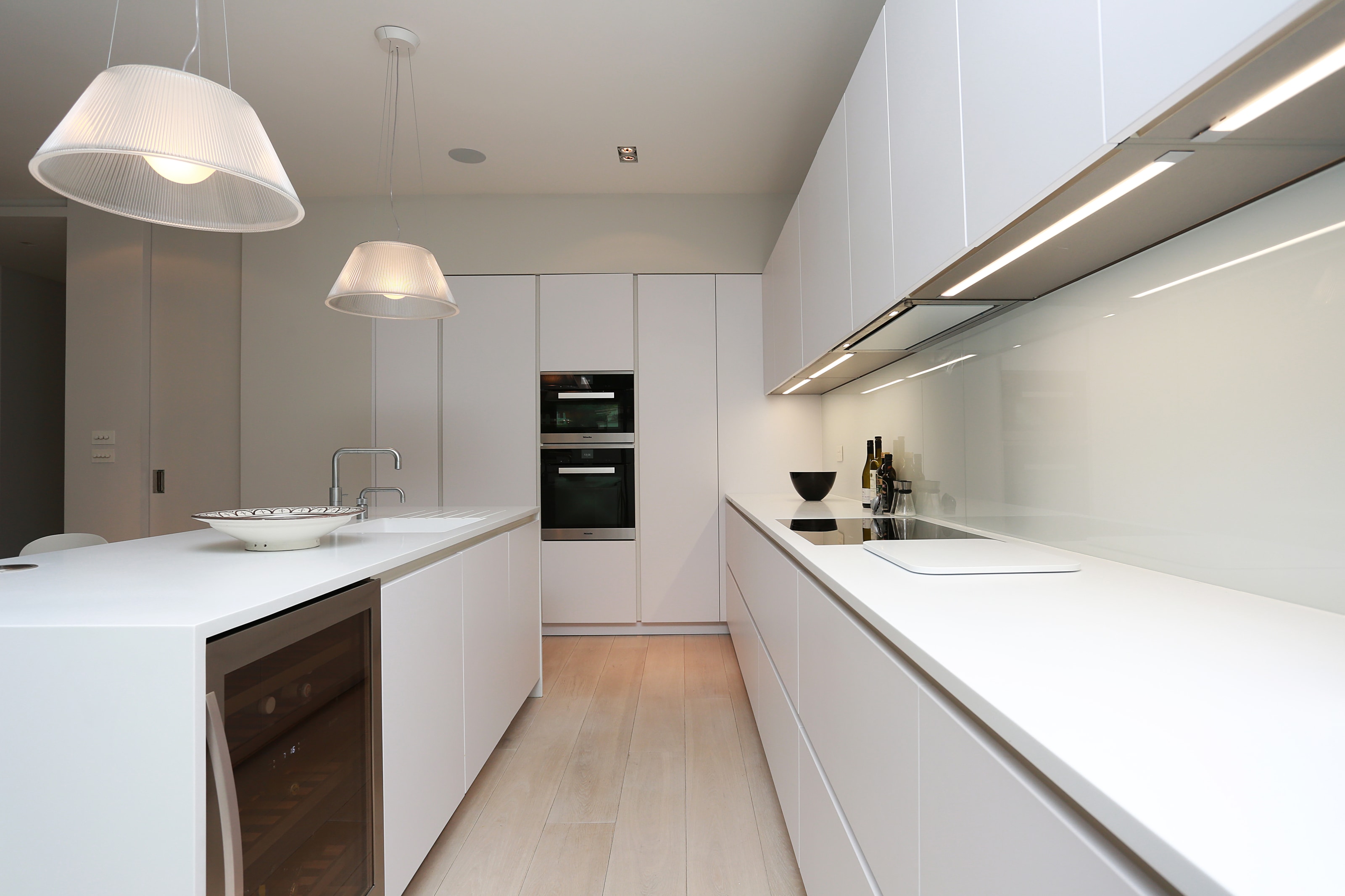 Hacker handleless kitchen in polar white with clean, fresh white cabinetry, a slimline island with integrated wine cooler, built-in double ovens, and a long run with induction hob and glass splashback under integrated lighting.