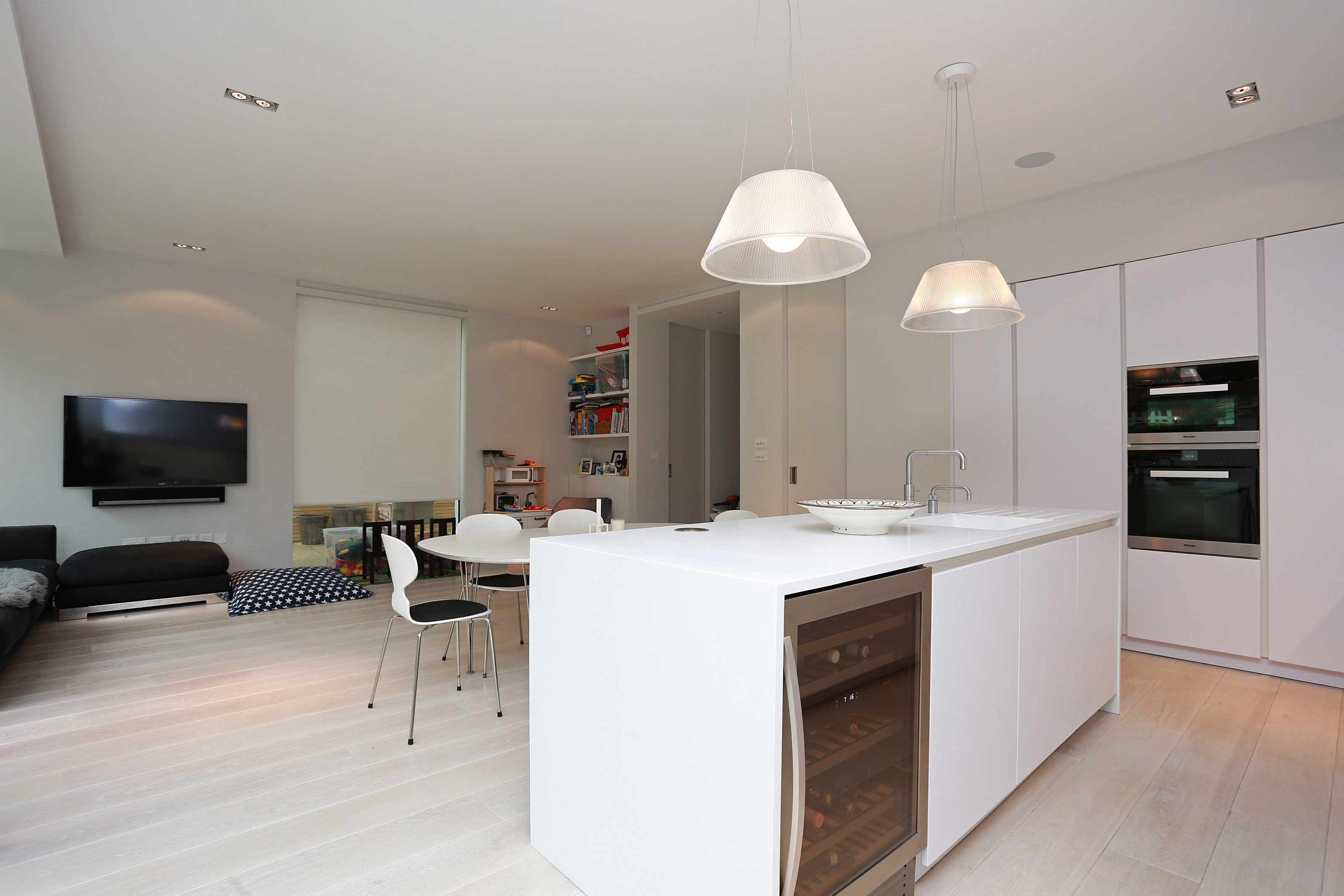 Open-plan Hacker kitchen in polar white with clean, fresh white handleless cabinetry, a large island with waterfall worktop, integrated sink and chrome tap, plus built-in ovens and an undercounter wine cooler opening onto a dining and living area.