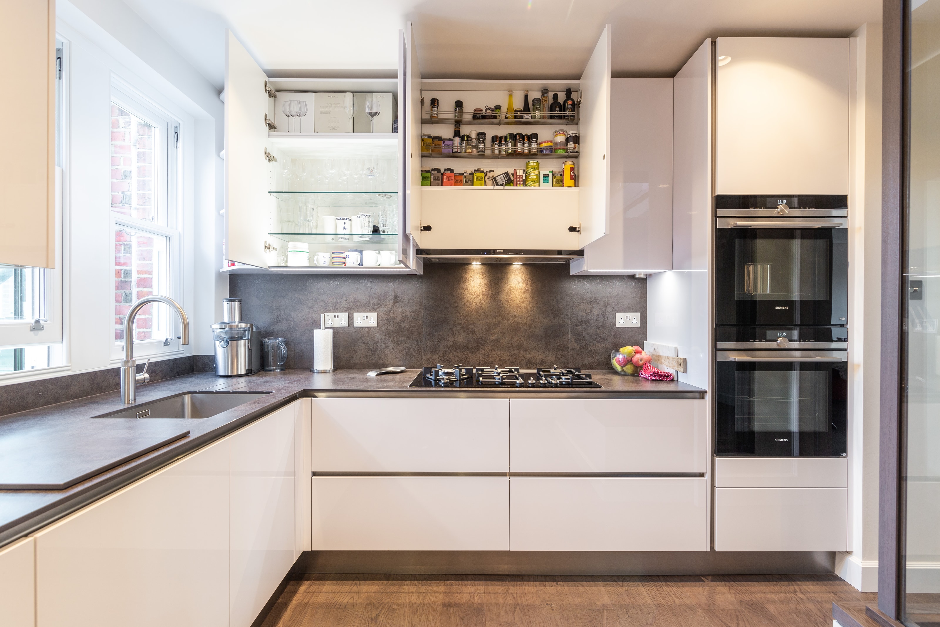 Compact L-shaped kitchen with high-gloss white handleless cabinets, dark stone-effect worktop and matching splashback, a stainless steel tap over an undermount sink by the window, a gas hob, and a tall housing with two built-in ovens; wall cupboards are open showing glass shelves and spice storage.