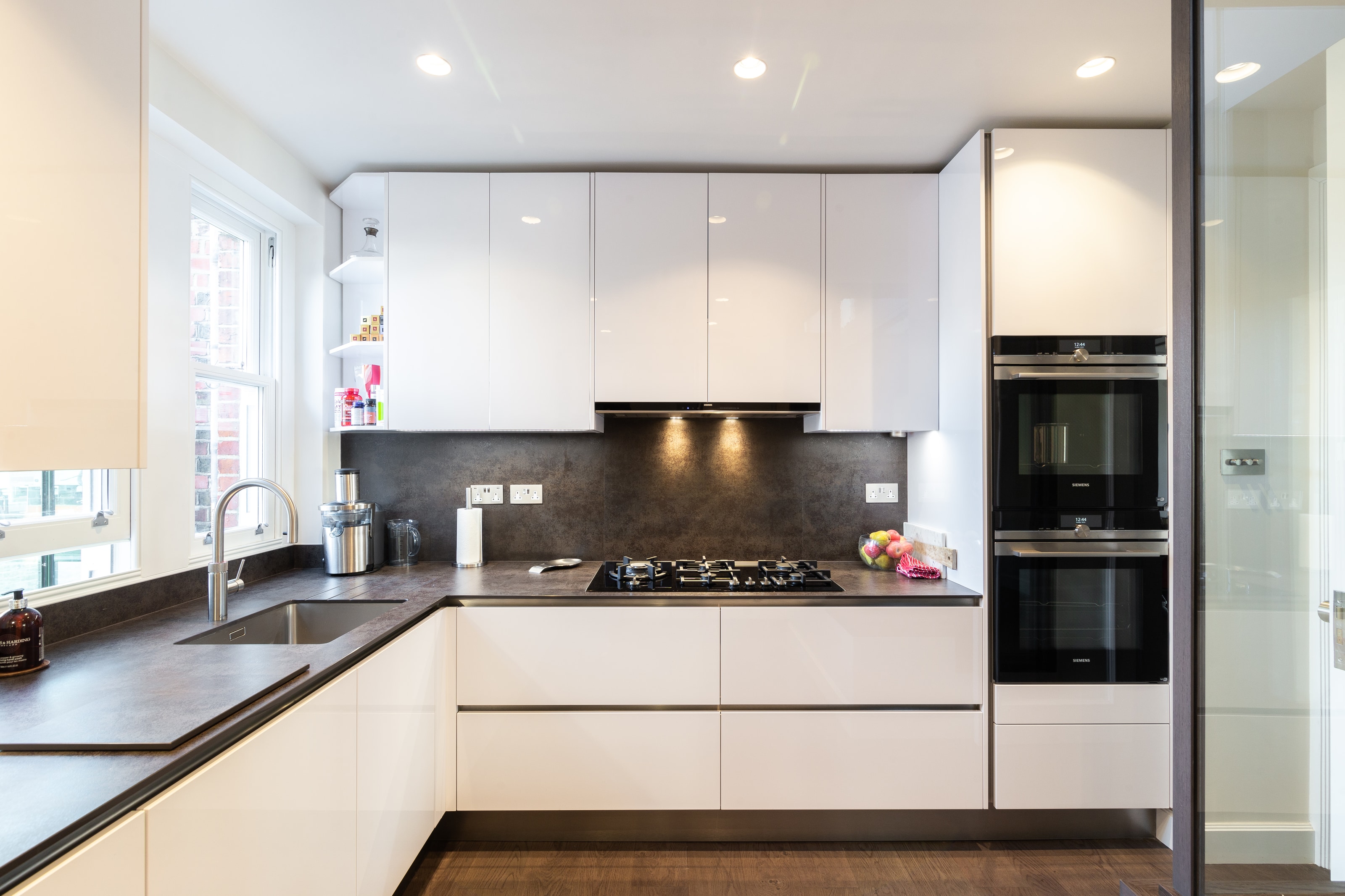 Modern L-shaped kitchen with gloss white handleless cabinets, a dark stone-effect worktop and matching splashback, undermount sink with chrome tap by a window, gas hob with integrated extractor lighting, and stacked black glass double ovens in a tall housing.