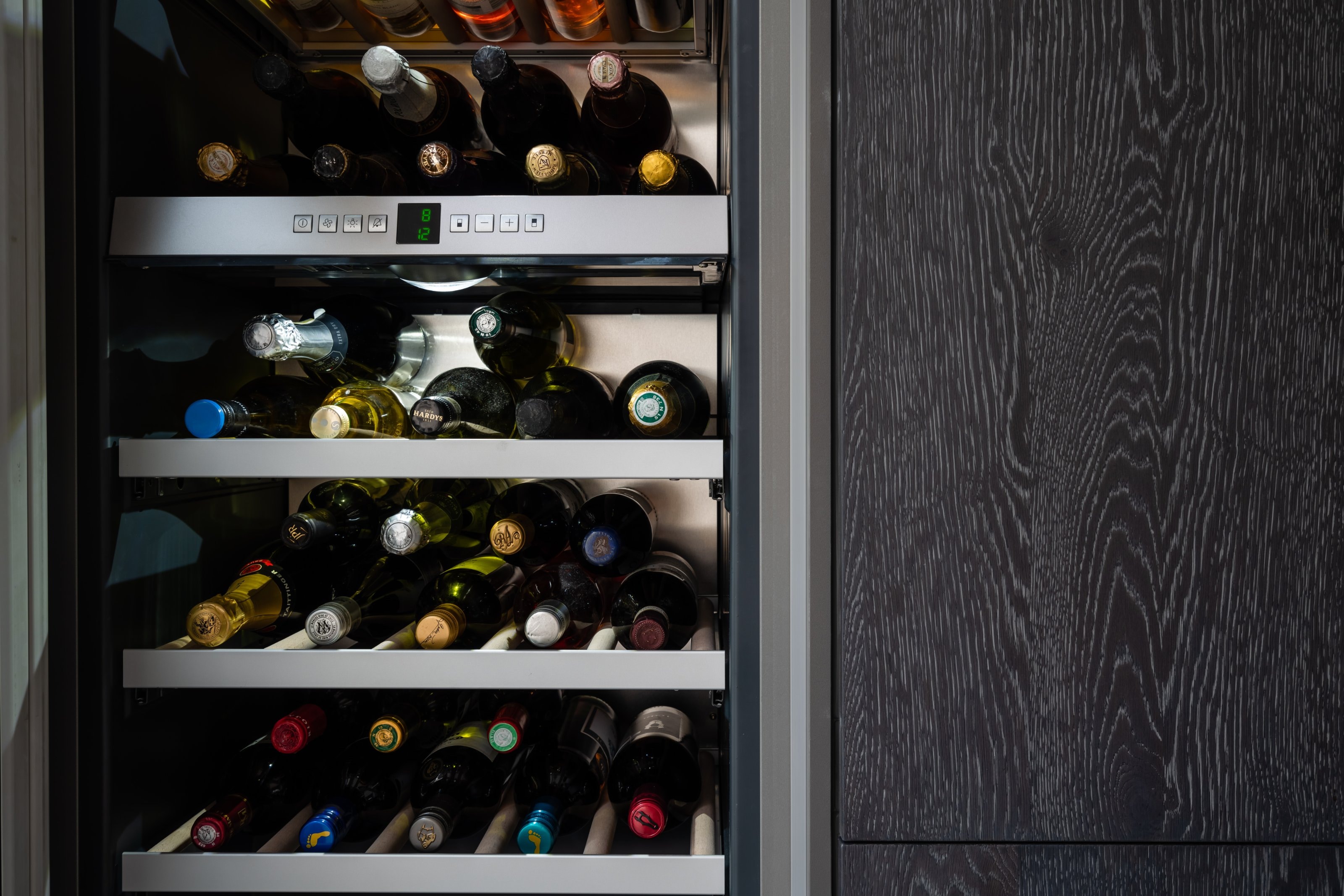 Close-up of a built-in wine cooler filled with bottles, set within textured dark wood kitchen cabinetry from Hacker.