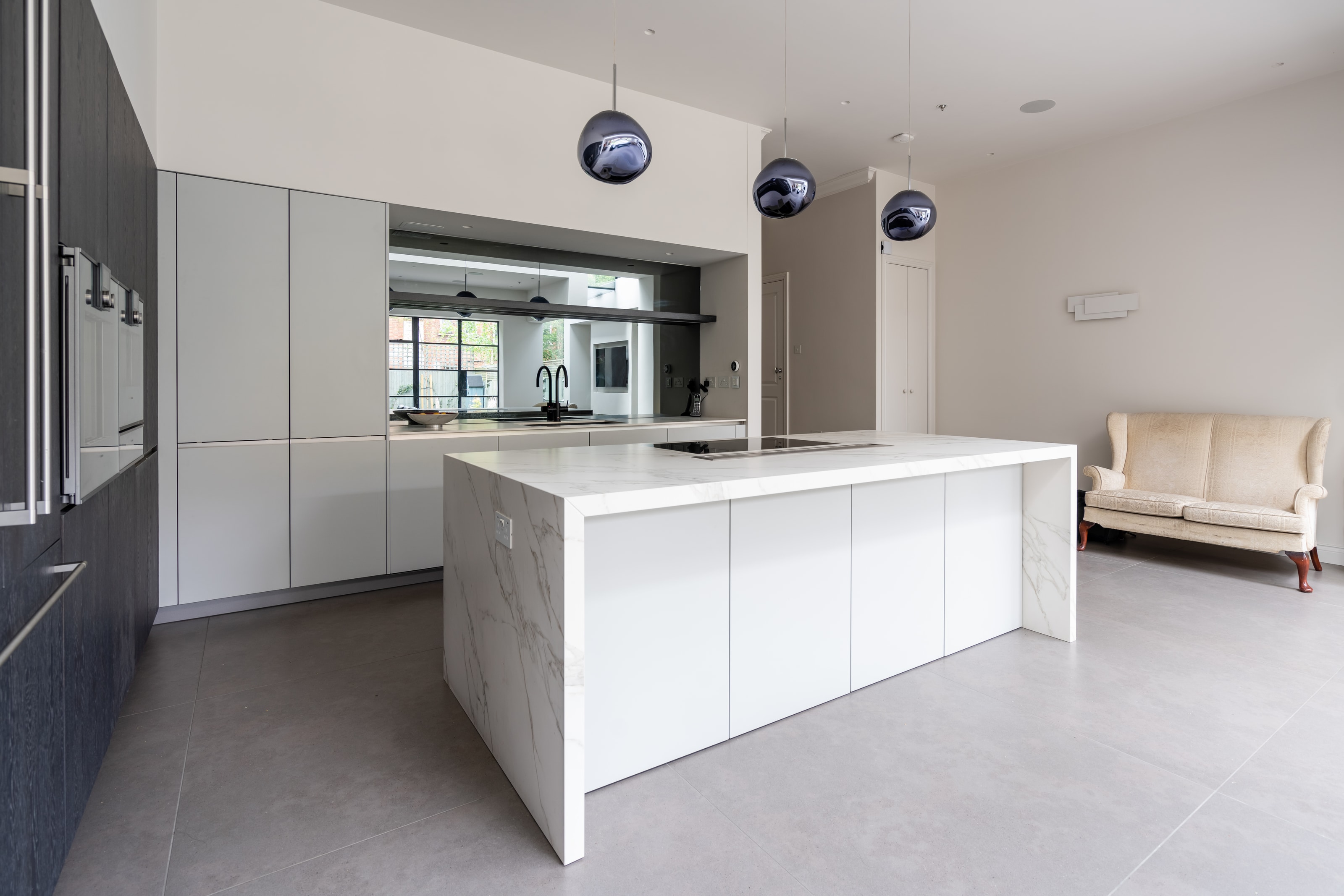 Open-plan Hacker kitchen with dark wood tall units and integrated Gaggenau appliances, paired with a white handleless island wrapped in marble-effect worktop and matching full-height sides, plus a black tap and grey tiled floor.