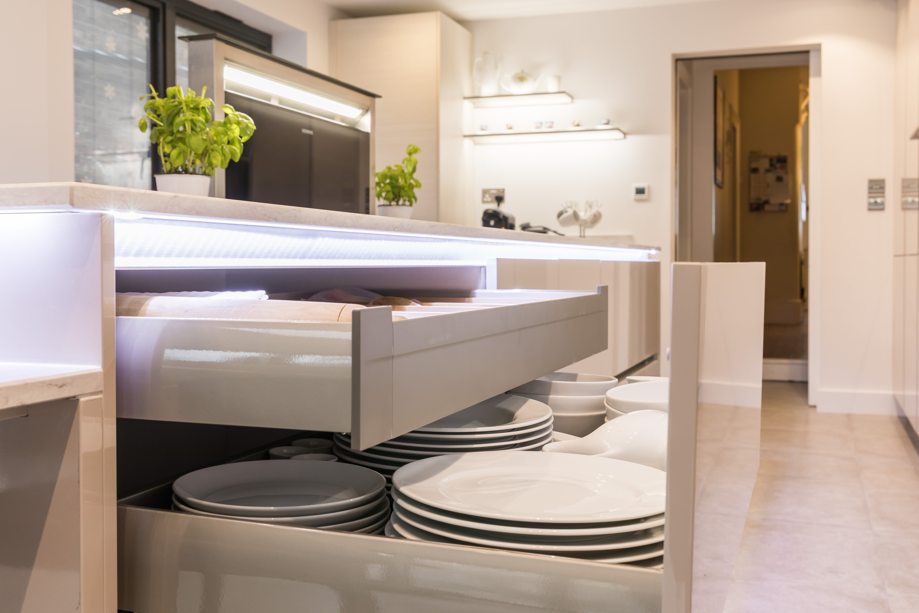 Open deep pan drawer in a Hacker handleless kitchen with cashmere gloss lacquer cabinetry, integrated LED drawer lighting and stacked white plates beneath a pale stone-effect worktop.