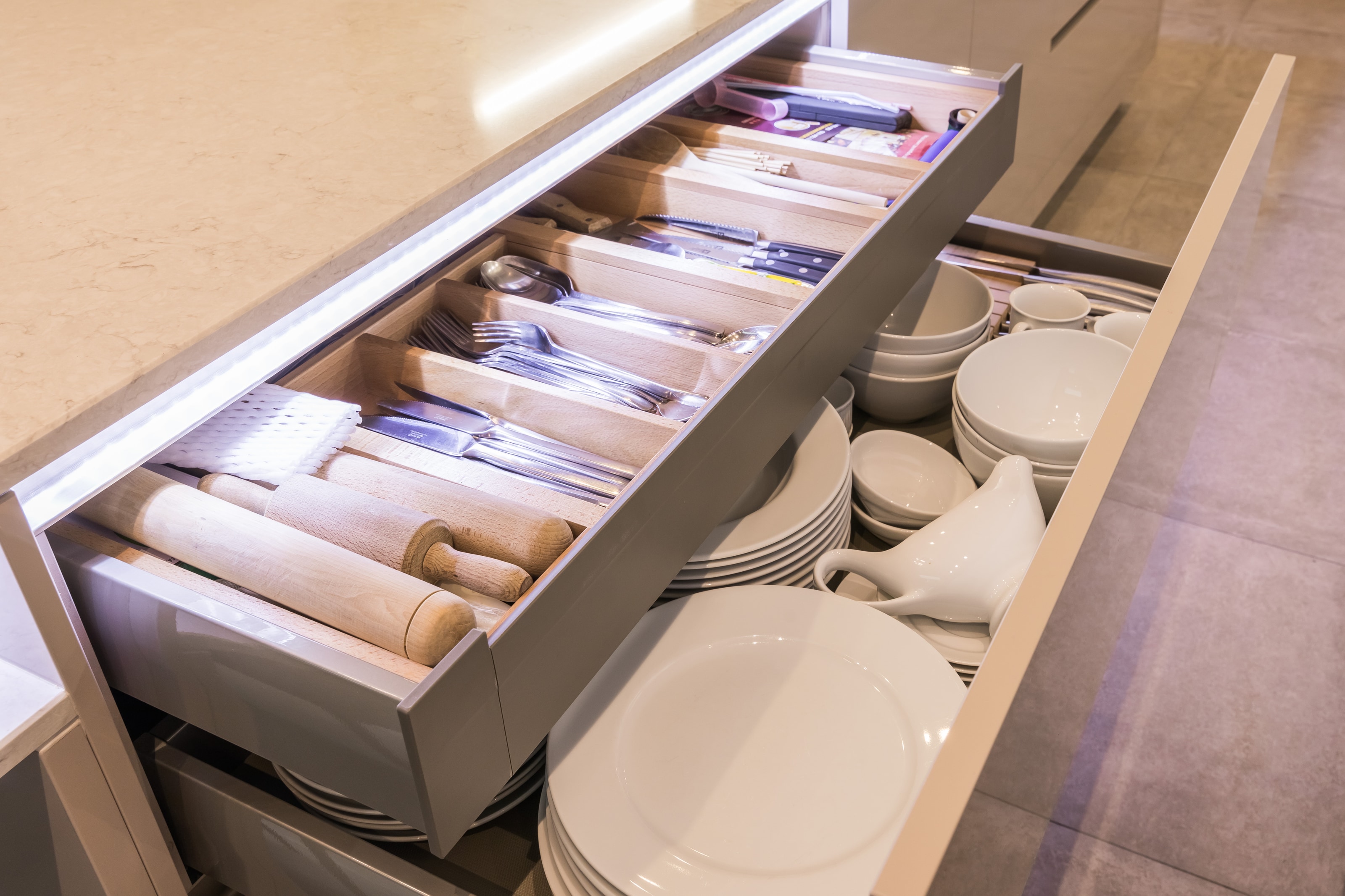 Open gloss kitchen drawers showing a wooden cutlery organiser with knives and utensils, and a deep drawer storing stacked white plates and bowls beneath a light stone-effect worktop.