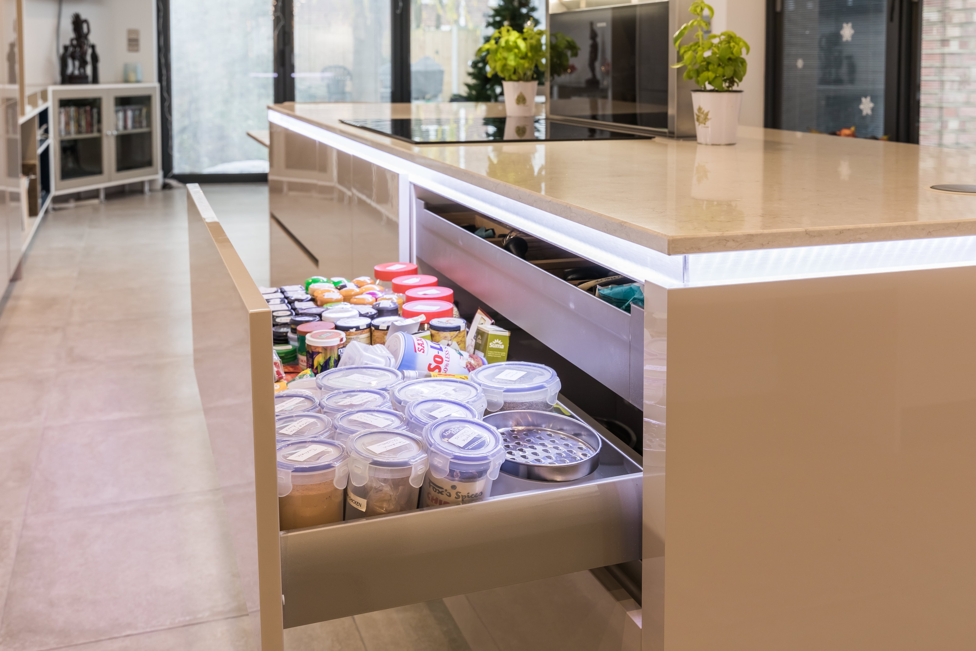 Hacker cashmere gloss lacquer kitchen island with a pale stone-effect worktop and flush hob, shown with a wide open deep drawer storing spices and containers beneath integrated under-worktop lighting.