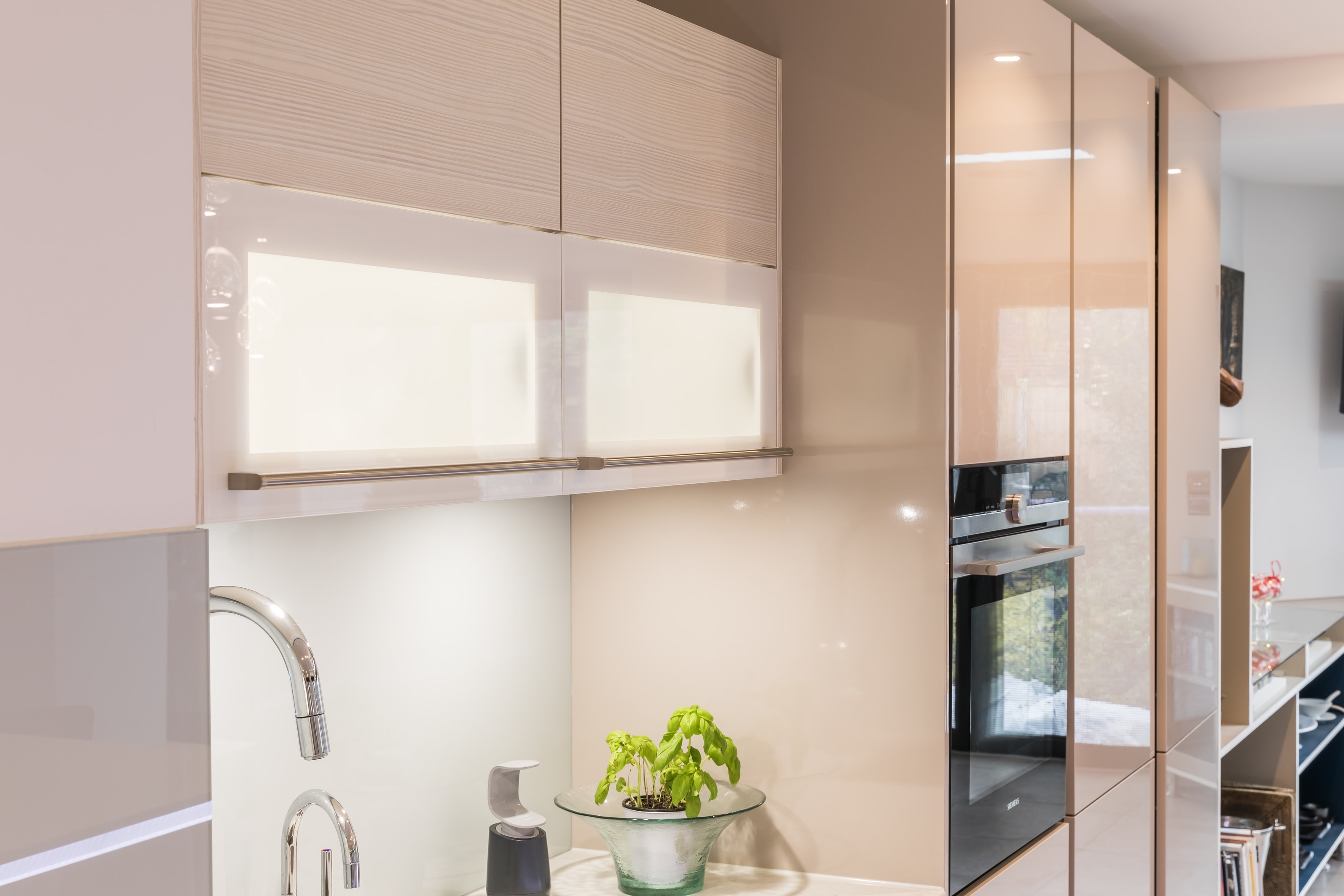 Detail of a Hacker kitchen in cashmere gloss lacquer with handleless wall units, a chrome mixer tap at the sink, and a built-in Siemens oven from the iq700 range beside a pale gloss splashback.