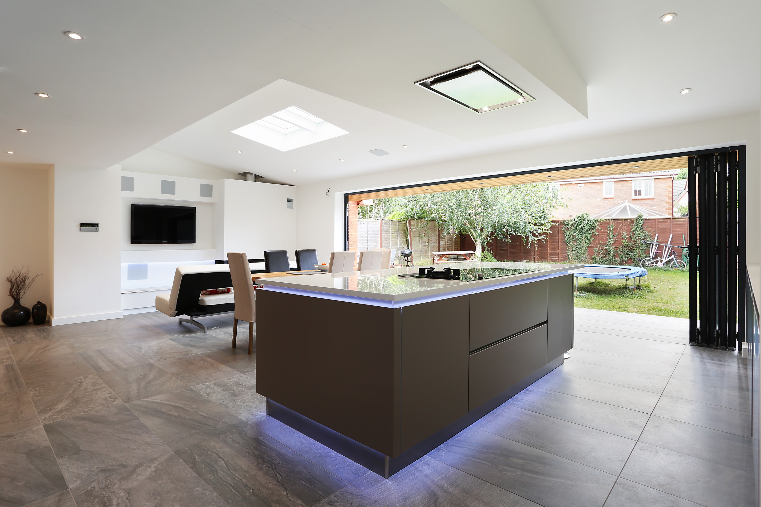 Hacker open-plan modern kitchen with a large two tone handleless island and breakfast bar, topped with a glossy stone-effect worktop and a flush hob, set on large-format grey floor tiles beside wide sliding doors to the garden.