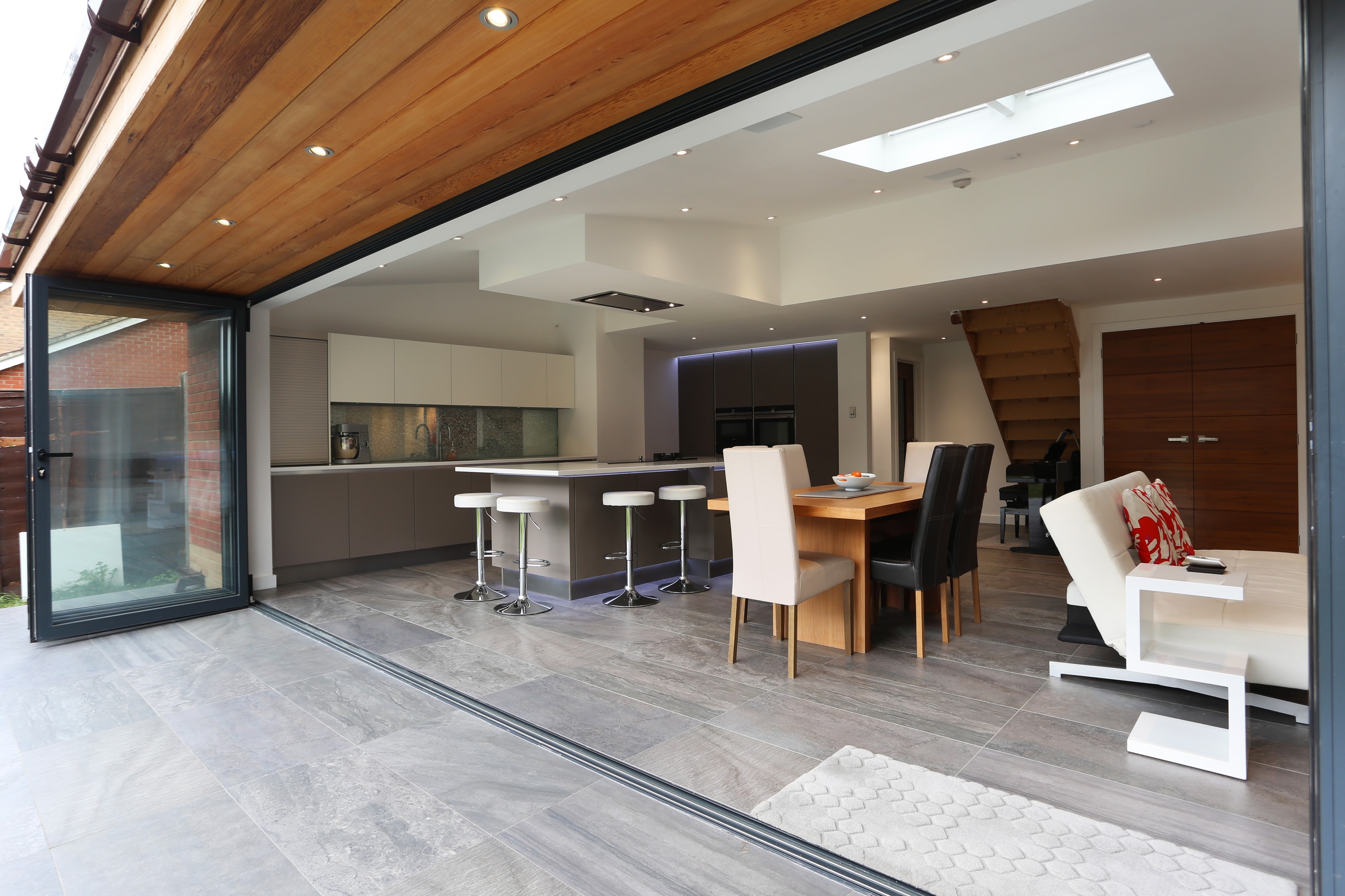 Open-plan modern Hacker kitchen with two-tone handleless cabinetry, a white breakfast bar island with chrome stools, grey floor tiles and a dining table beside large sliding doors.
