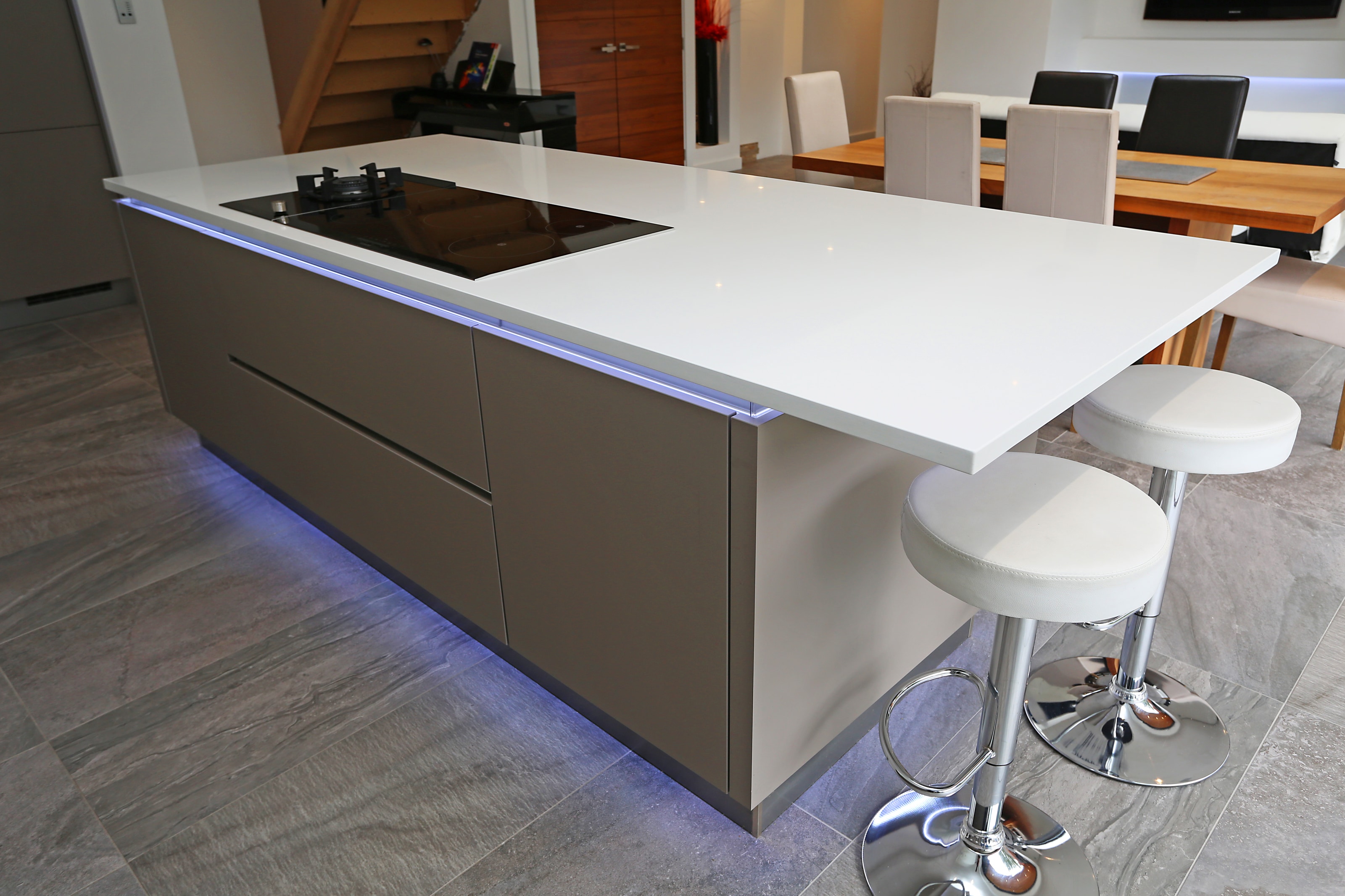 Modern Hacker open-plan kitchen with a two tone handleless island in matt grey and white, a white worktop with integrated hob, blue plinth lighting and a breakfast bar with white stools beside the dining area.