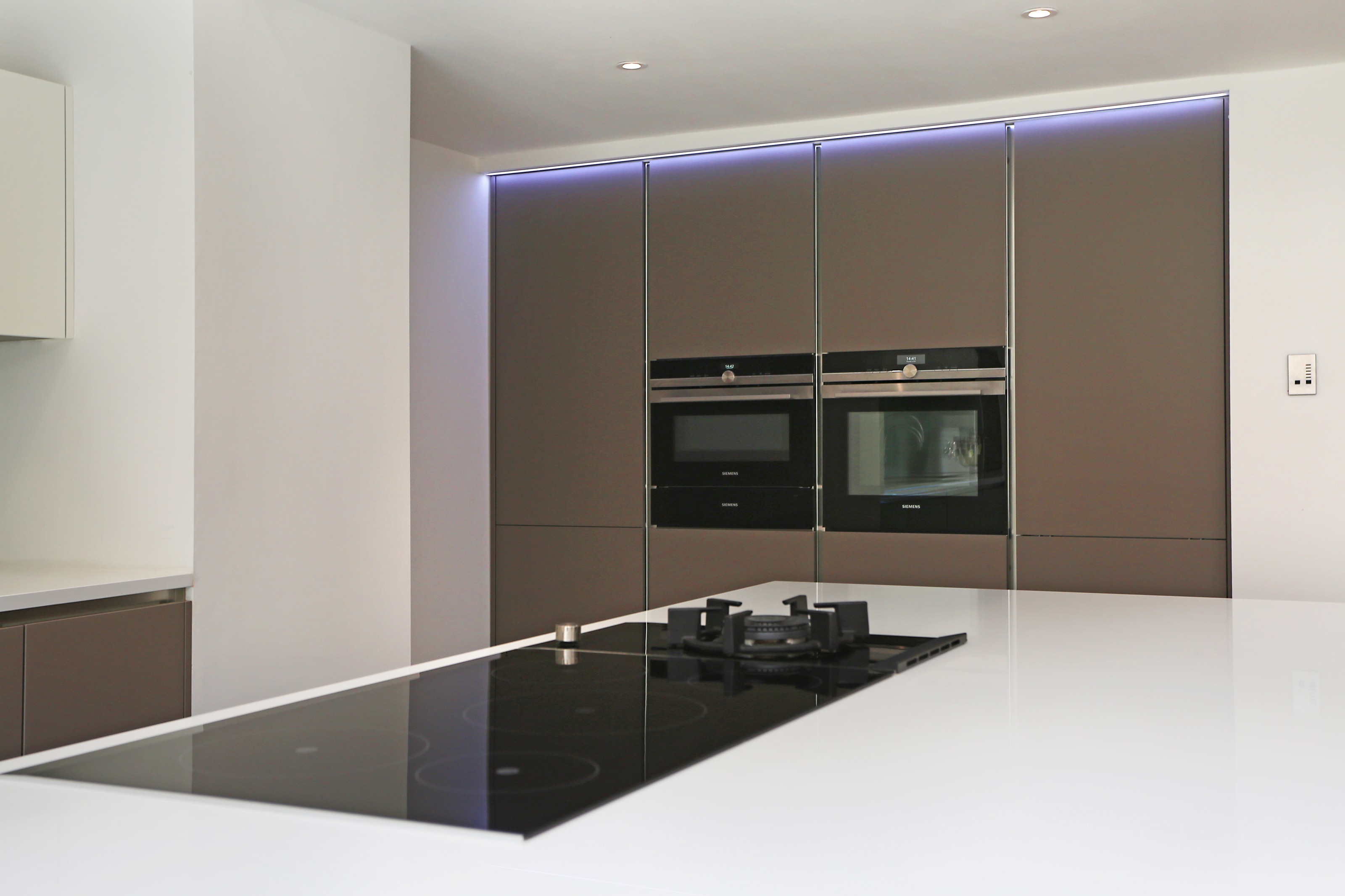 Modern Hacker kitchen in a two tone scheme, with a large white breakfast bar island and black induction hob, facing tall matt taupe handleless cabinetry with built-in ovens and subtle LED lighting in an open plan space.