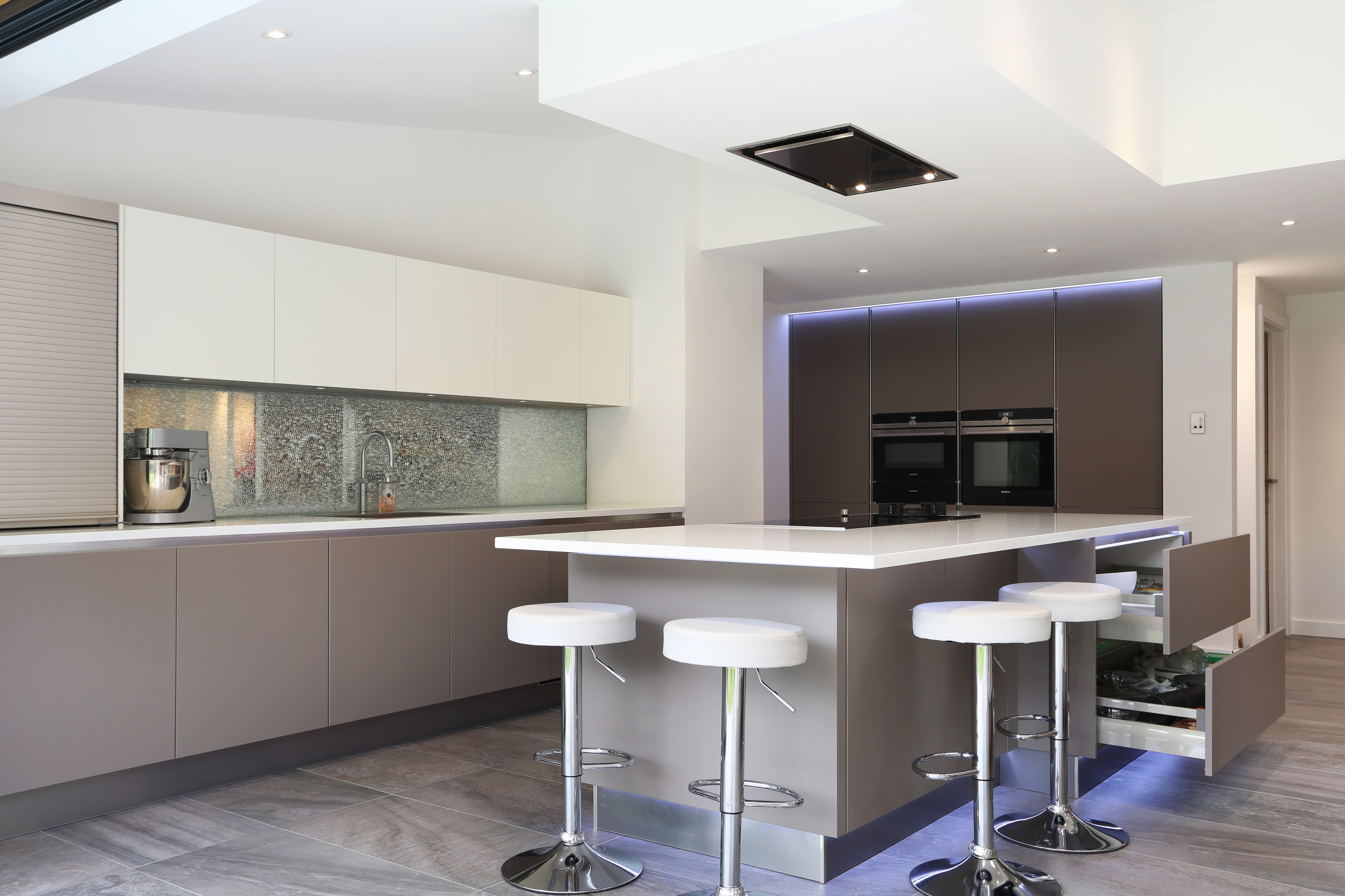 Modern Hacker two-tone open-plan kitchen with a white breakfast bar island, matt taupe handleless cabinetry, white wall units, mirrored splashback and built-in ovens, with chrome bar stools and grey tiled flooring.