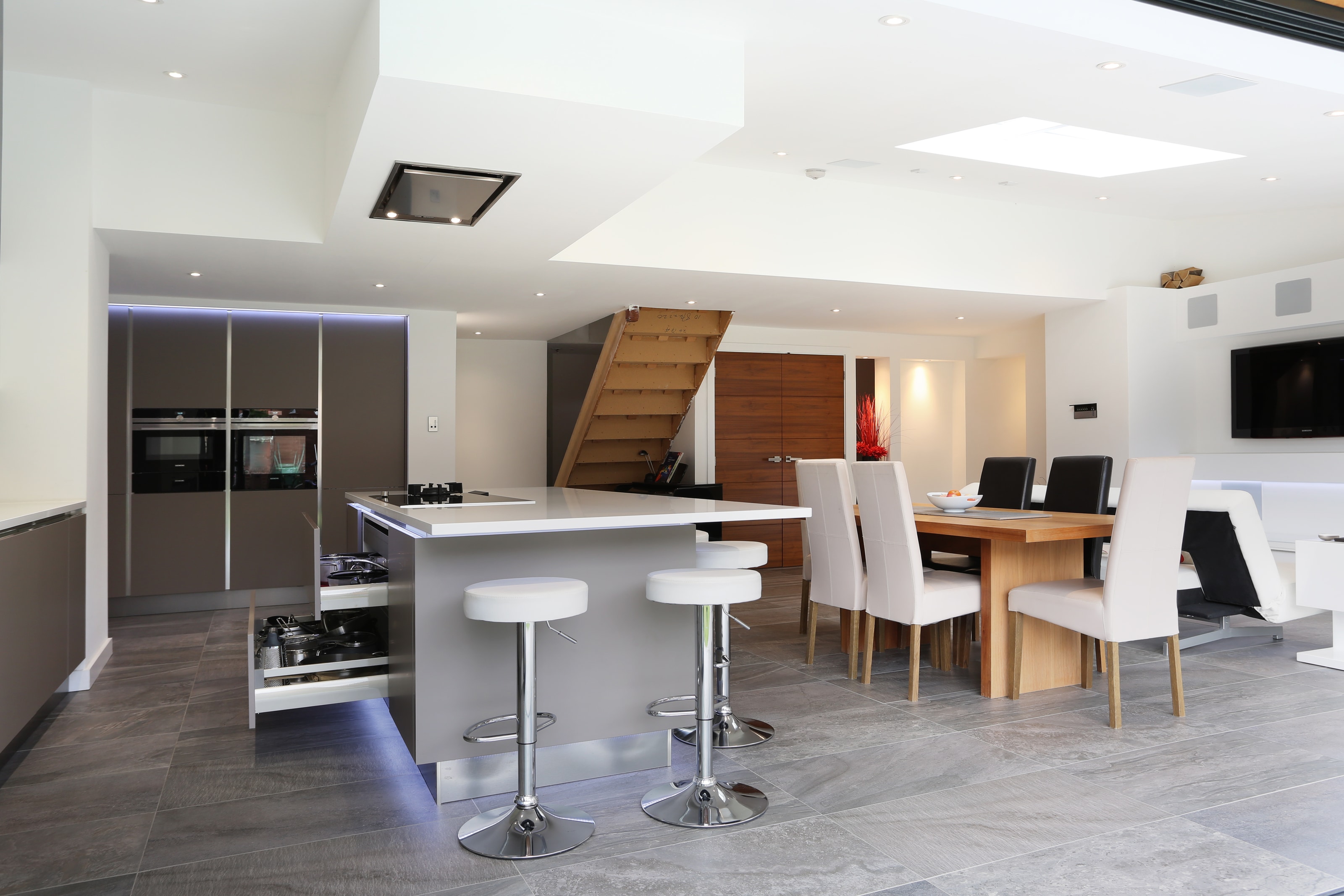 Modern open plan Hacker kitchen in two tone grey and white, with a large island and breakfast bar stools, white worktop, integrated ovens, and an open dining area with a wooden table and upholstered chairs.