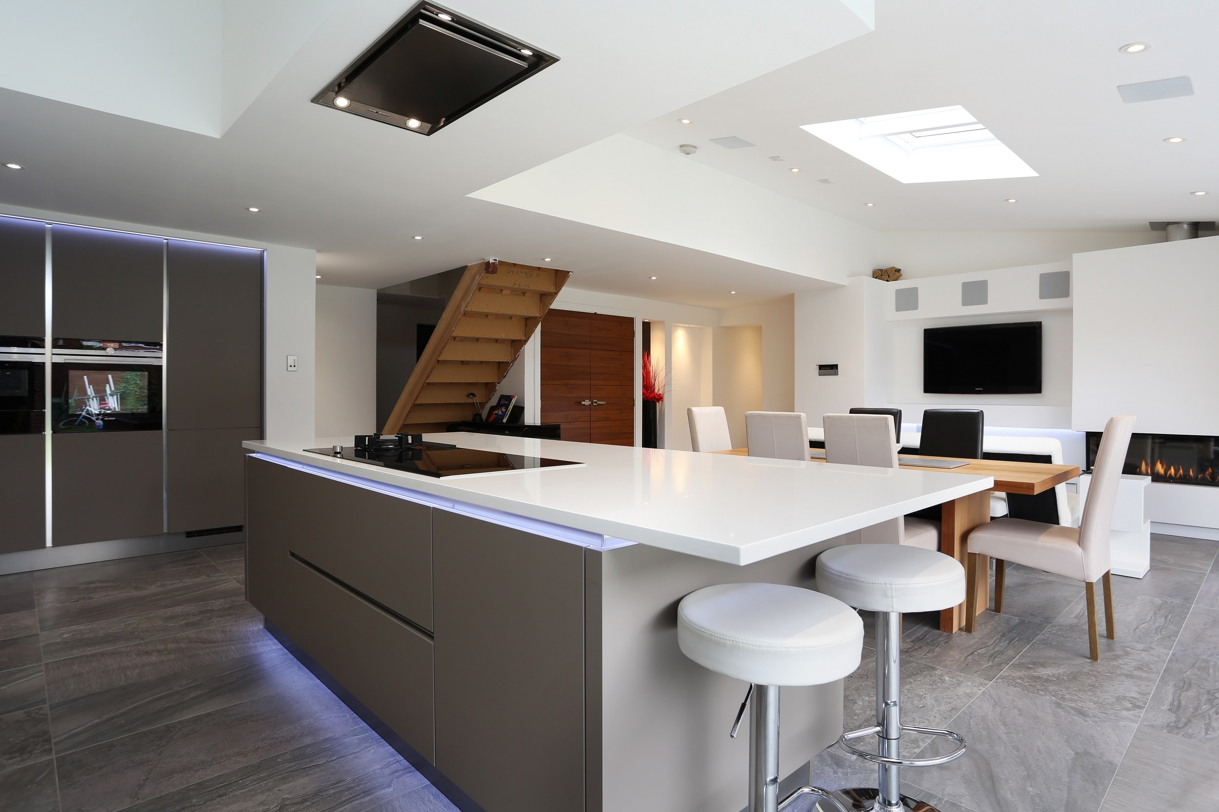 Open-plan modern Hacker kitchen with two tone handleless grey cabinetry, a large white island with breakfast bar seating, integrated ovens, and recessed lighting leading into a dining area and media wall.