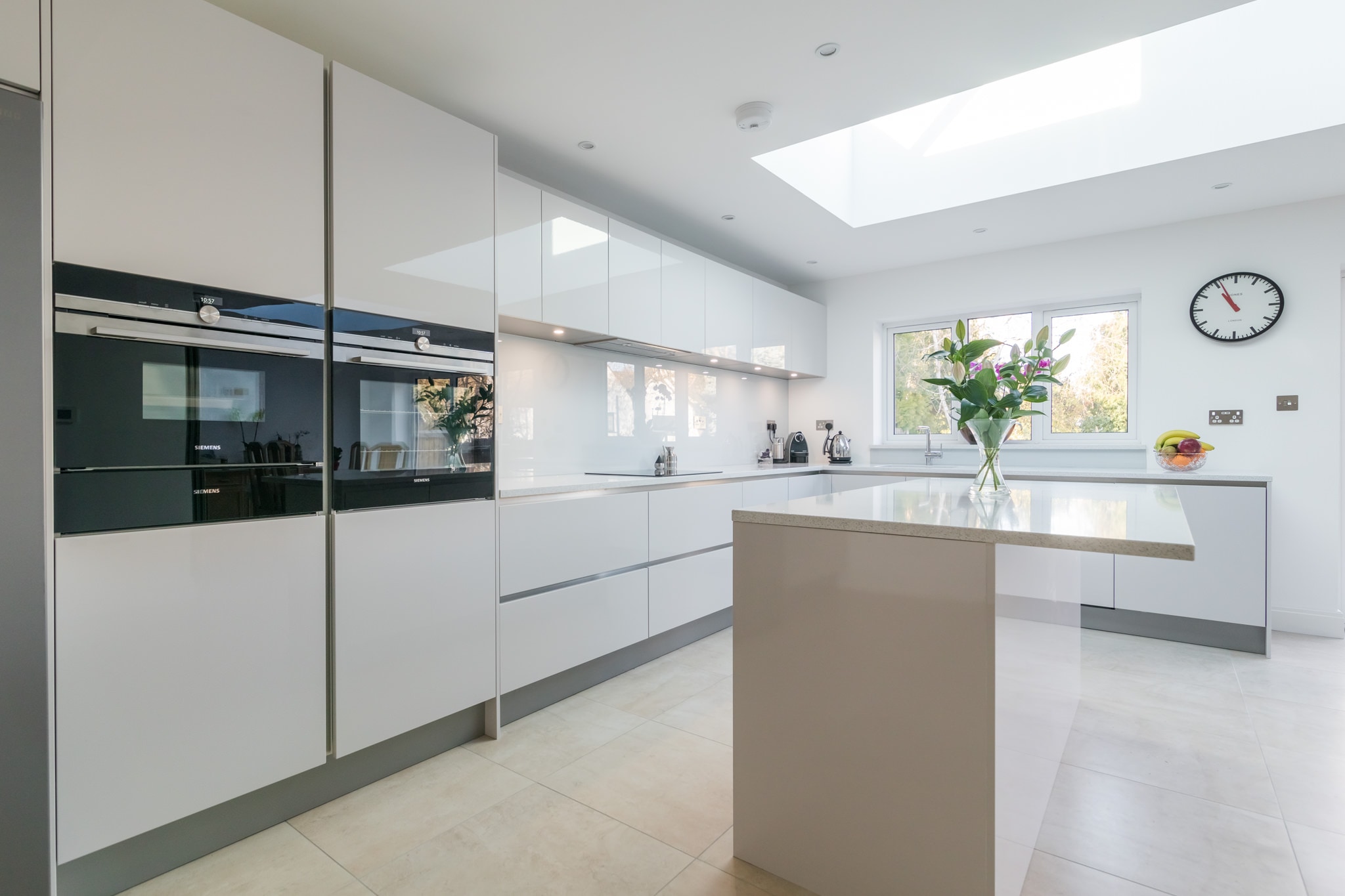 Contemporary Hacker handleless kitchen with high-gloss taupe and white cabinetry, Siemens built-in ovens, an L-shaped run and a matching island with a light stone-effect worktop under a large rooflight.