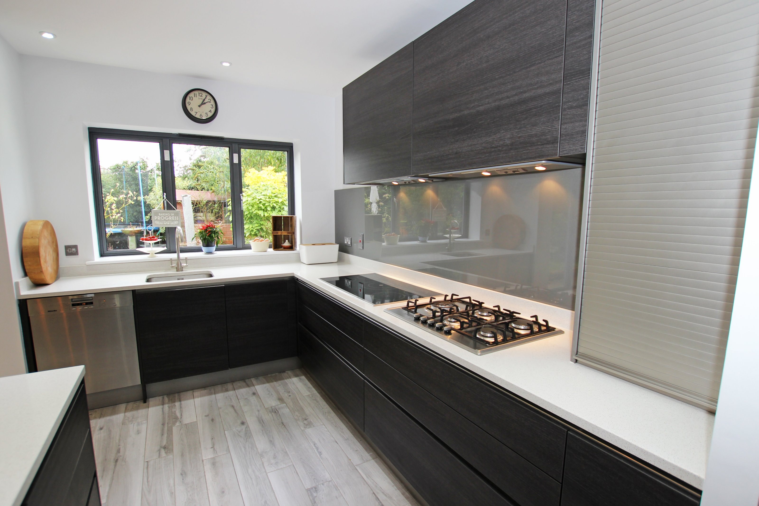 Contemporary Hacker kitchen with dark wood-effect handleless units, pale worktops and a grey glass splashback, featuring a gas hob run and an under-window sink with stainless steel dishwasher.