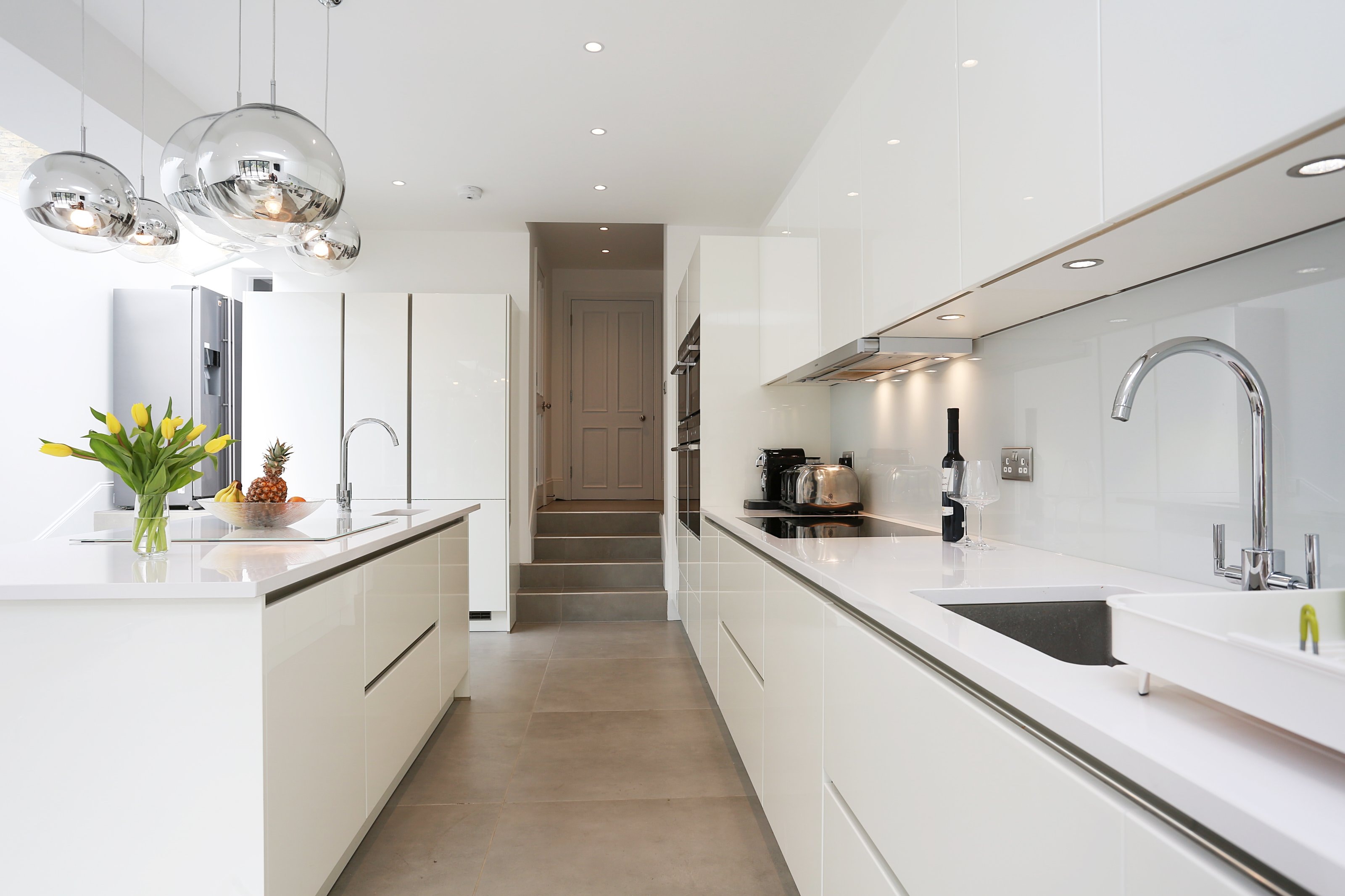 Bright open-plan Hacker kitchen with gloss white handleless cabinetry, a white island breakfast bar with inset hob and sink, long white worktops, chrome taps, integrated ovens and large reflective pendant lights.