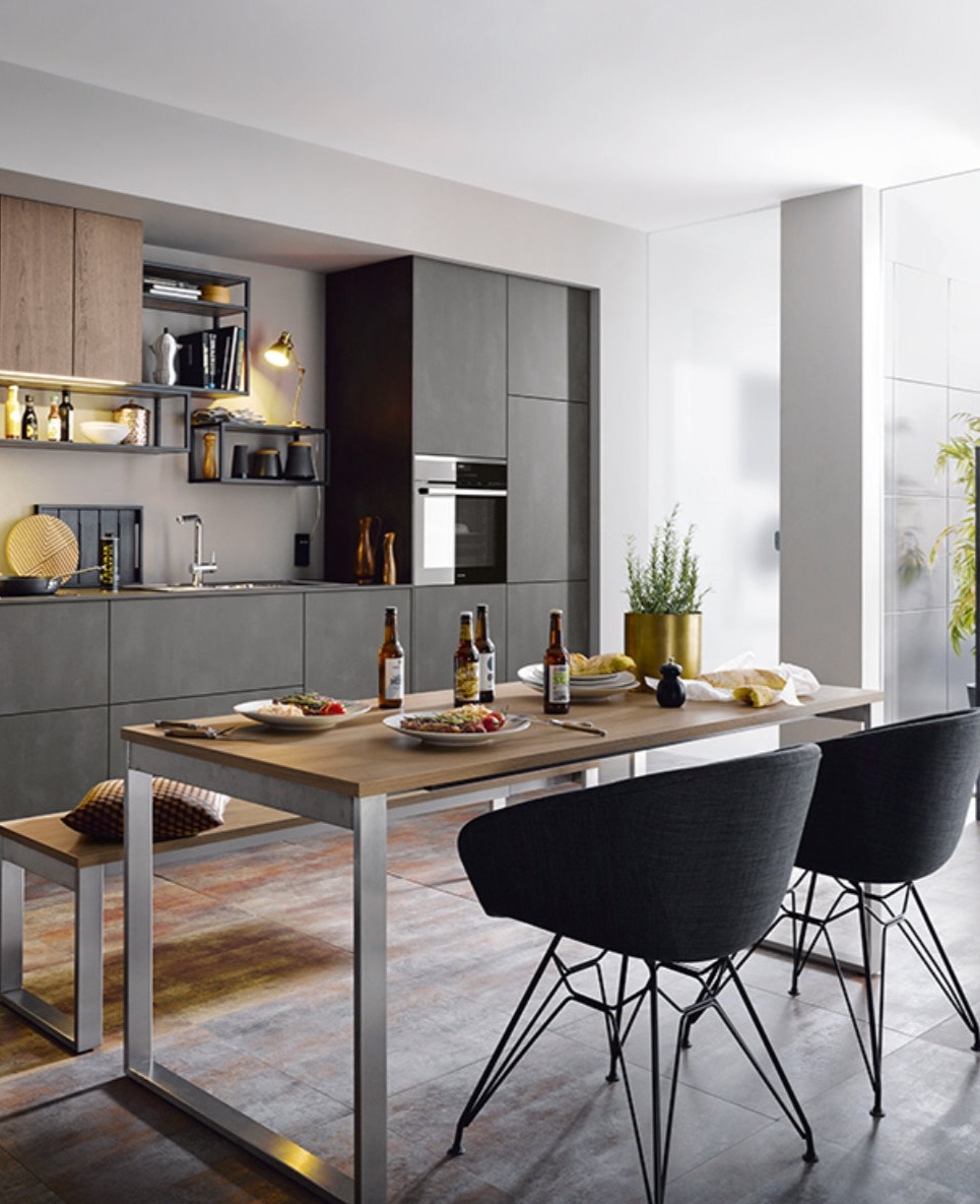 Open-plan Schuller kitchen with handleless grey ceramic cabinetry, wood wall units and a matching wood dining table, plus open shelving, integrated oven and a slimline chrome tap at the sink.