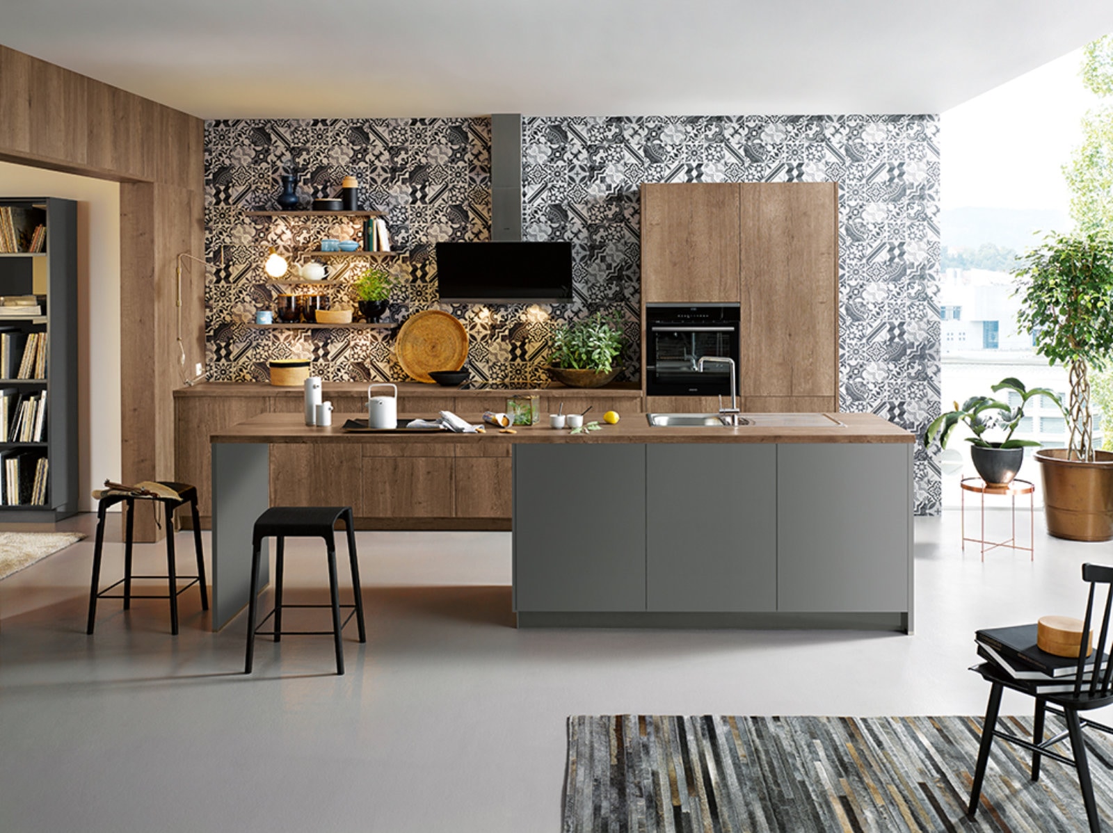 Open-plan Schuller Kitchens design with matt wood-effect handleless cabinetry, a large island with grey base units and a wood-effect worktop, integrated oven housing, and a sink set into the island. Patterned monochrome wall tiles, open shelving with styling, black bar stools and indoor plants complete the space.