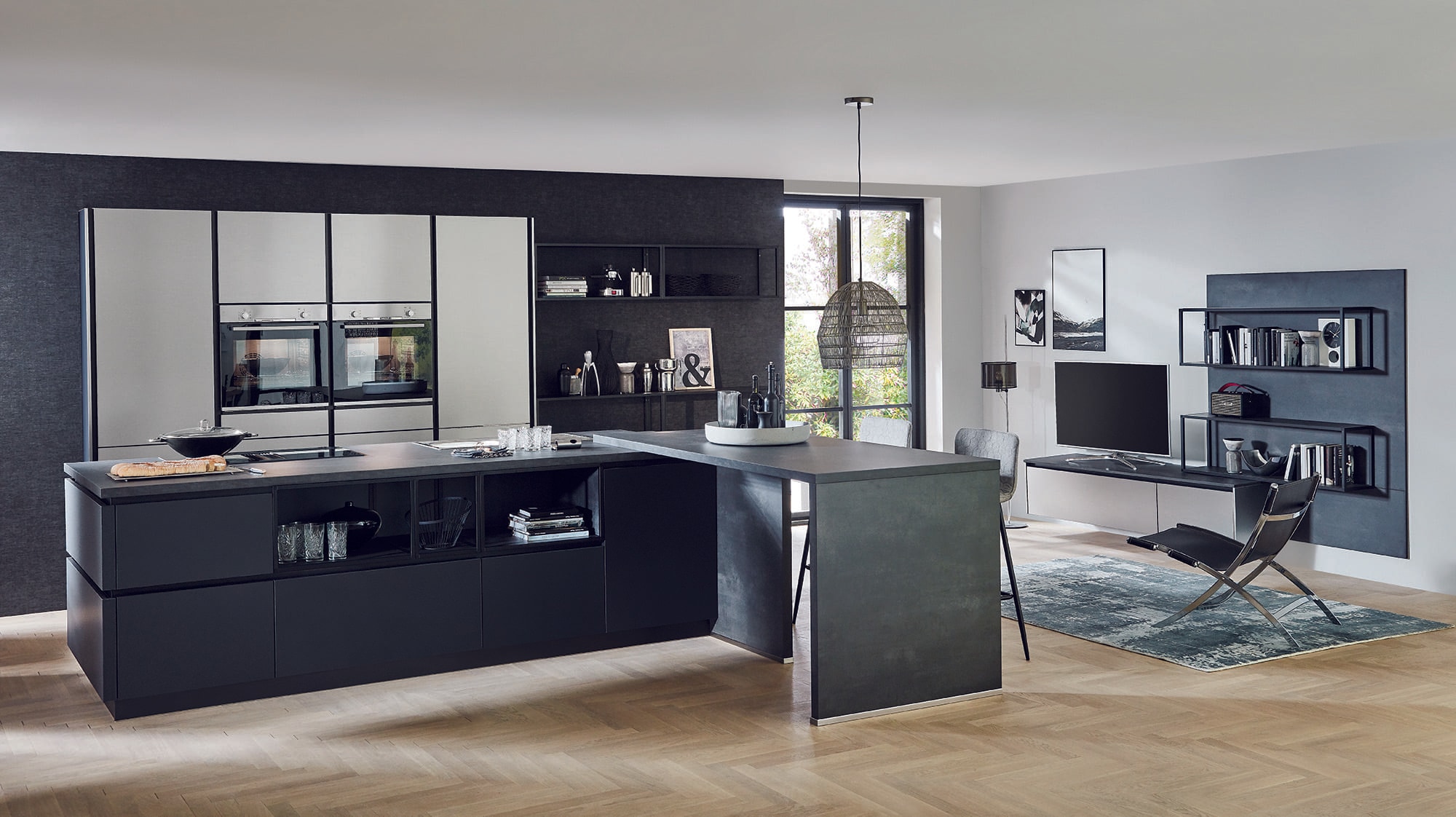 Nobilia Black Matt Metallic Handleless Kitchen with dark matt base units, a large island and peninsular breakfast bar, grey stone-effect worktops, and tall light cabinetry housing built-in ovens, flowing into a minimalist living area with wall shelving and a TV.