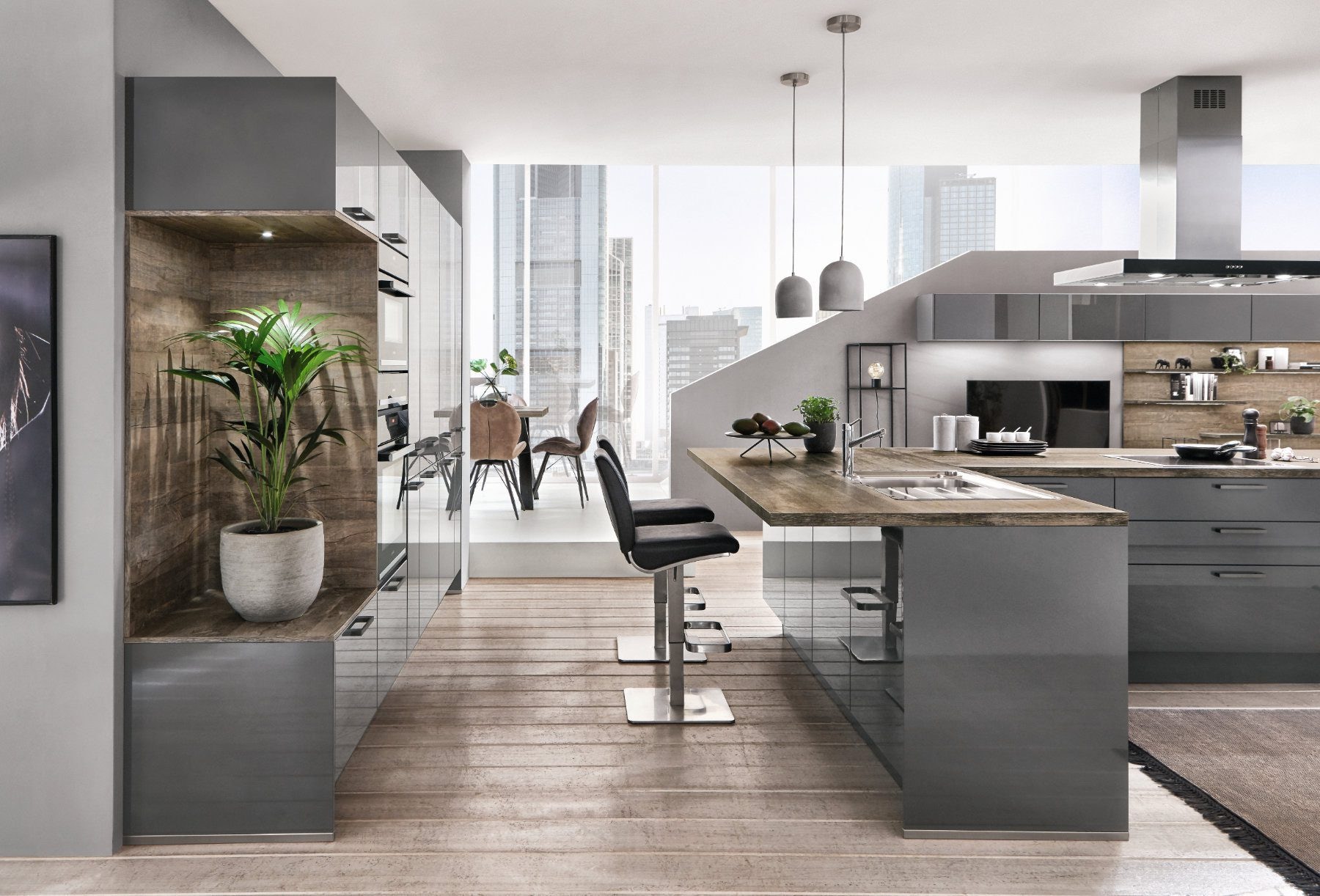 Open-plan Nobilia grey gloss kitchen with a peninsula breakfast bar, wood-effect worktop and splashback, built-in ovens, chrome tap and sink, and a stainless steel cooker hood, leading to a dining area by full-height windows.