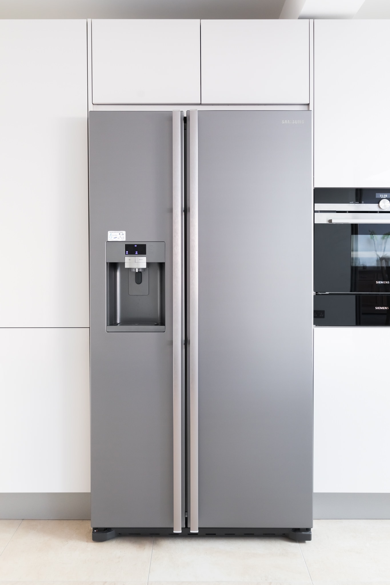 Stainless steel American-style fridge freezer with water dispenser, set within a minimalist handleless Hacker kitchen run with white gloss cabinetry and Siemens built-in ovens.